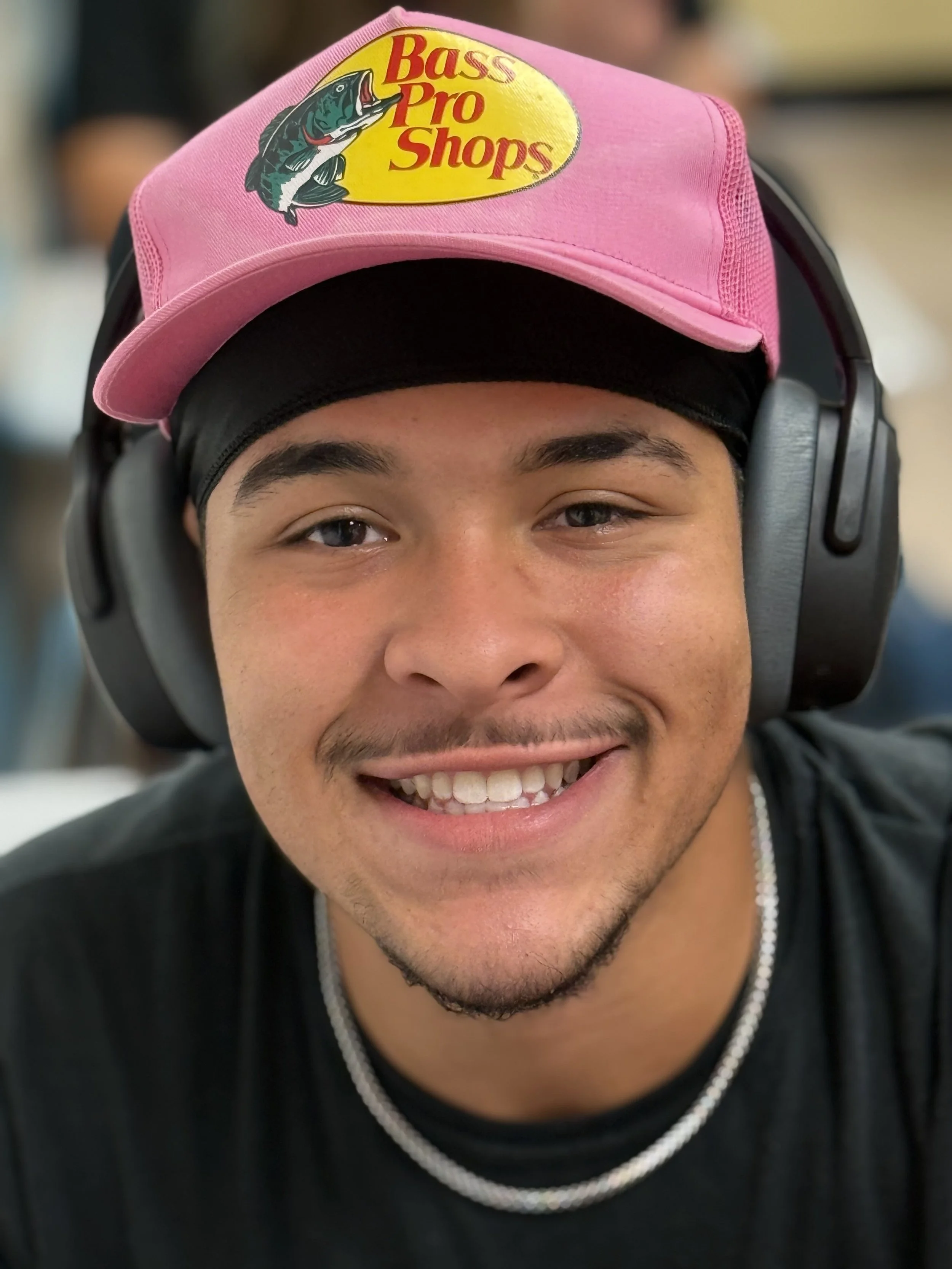 Close-up of a young man smiling, wearing a pink Bass Pro Shops cap, black head covering, black shirt, and headphones.