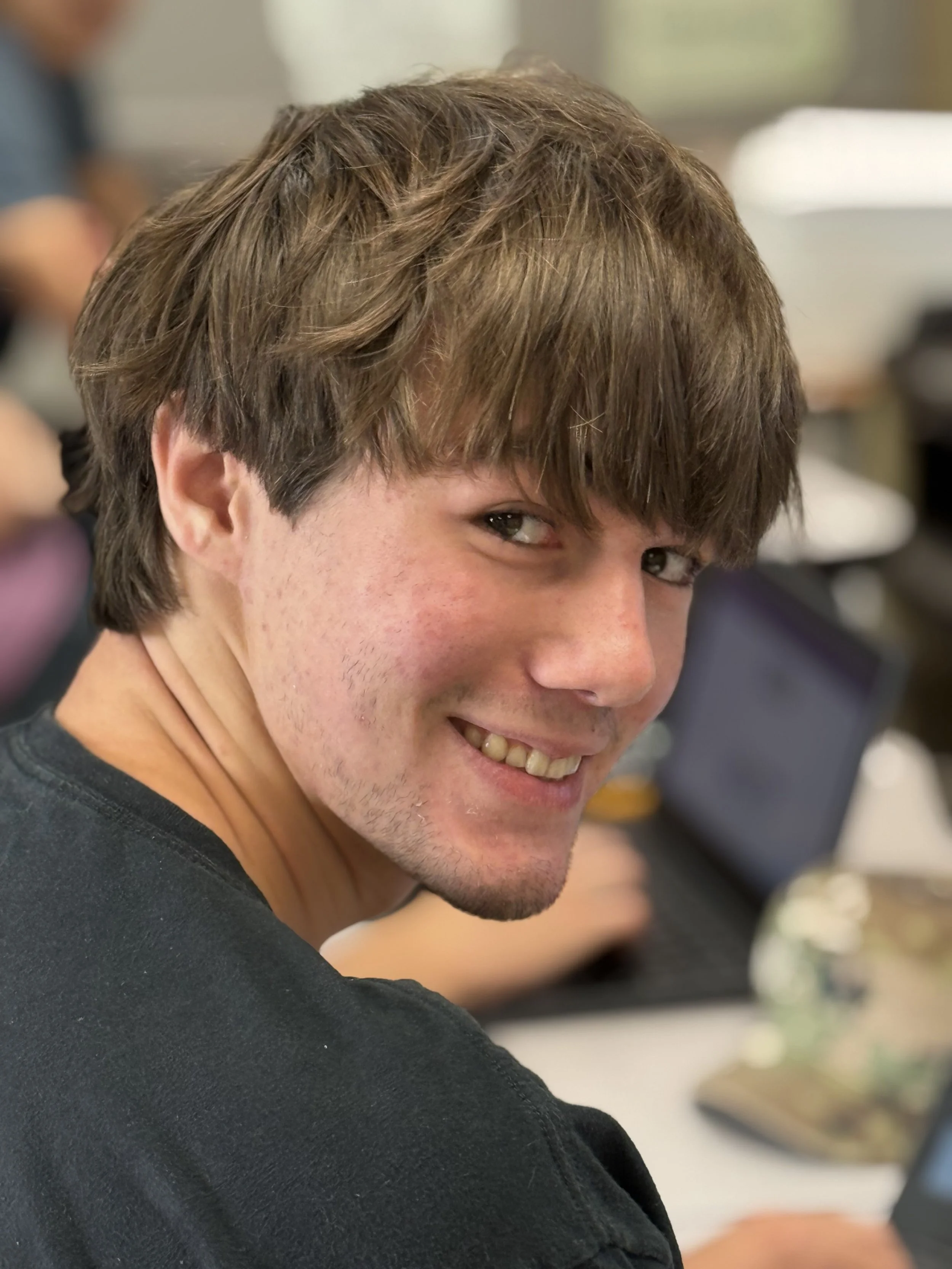 A young man with brown hair and a light beard smiling while looking over his shoulder in a classroom or office setting.