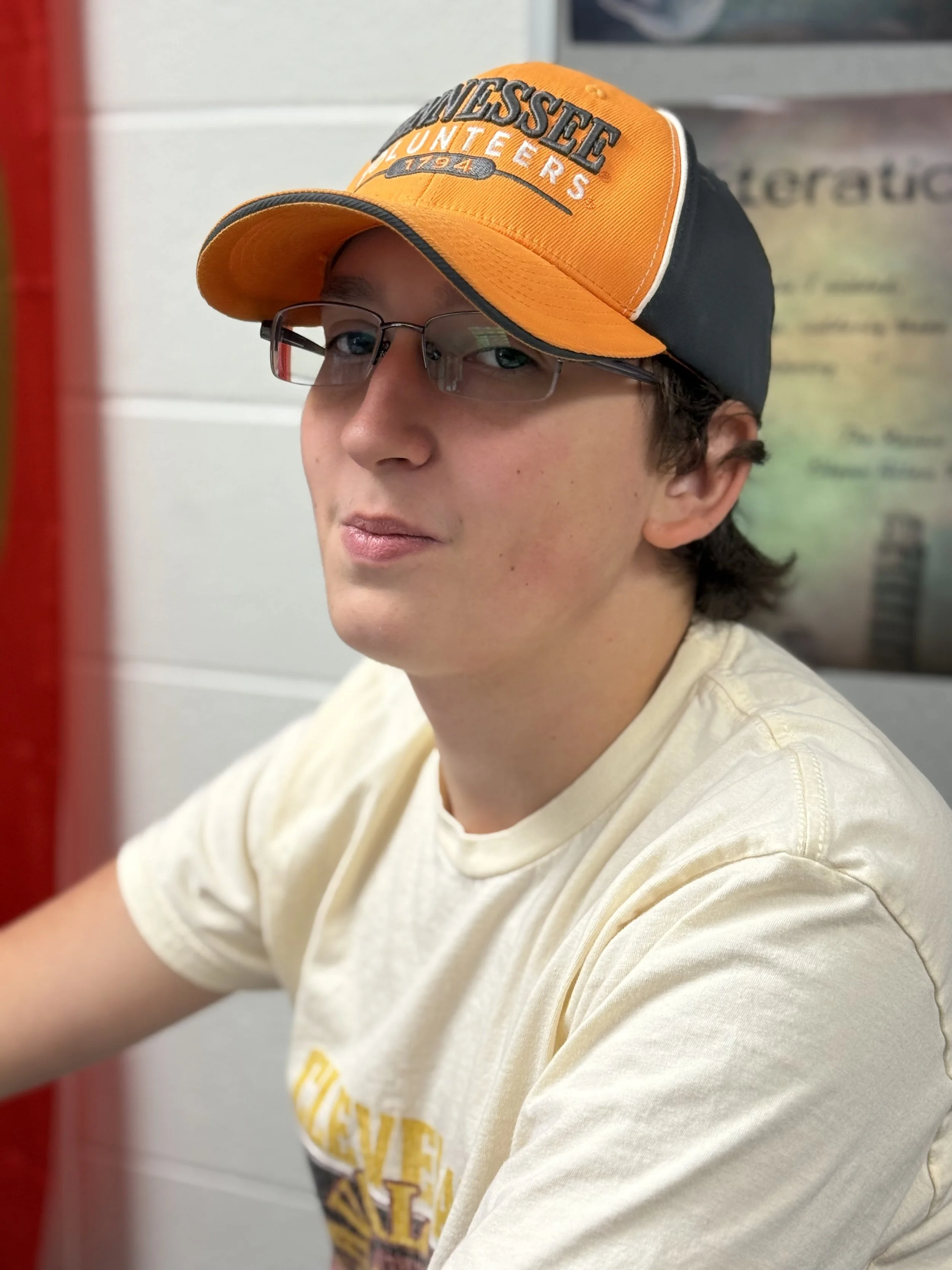 A young man wearing glasses, a Tennessee Volunteers cap, and a light-colored t-shirt sitting in front of a wall with posters.