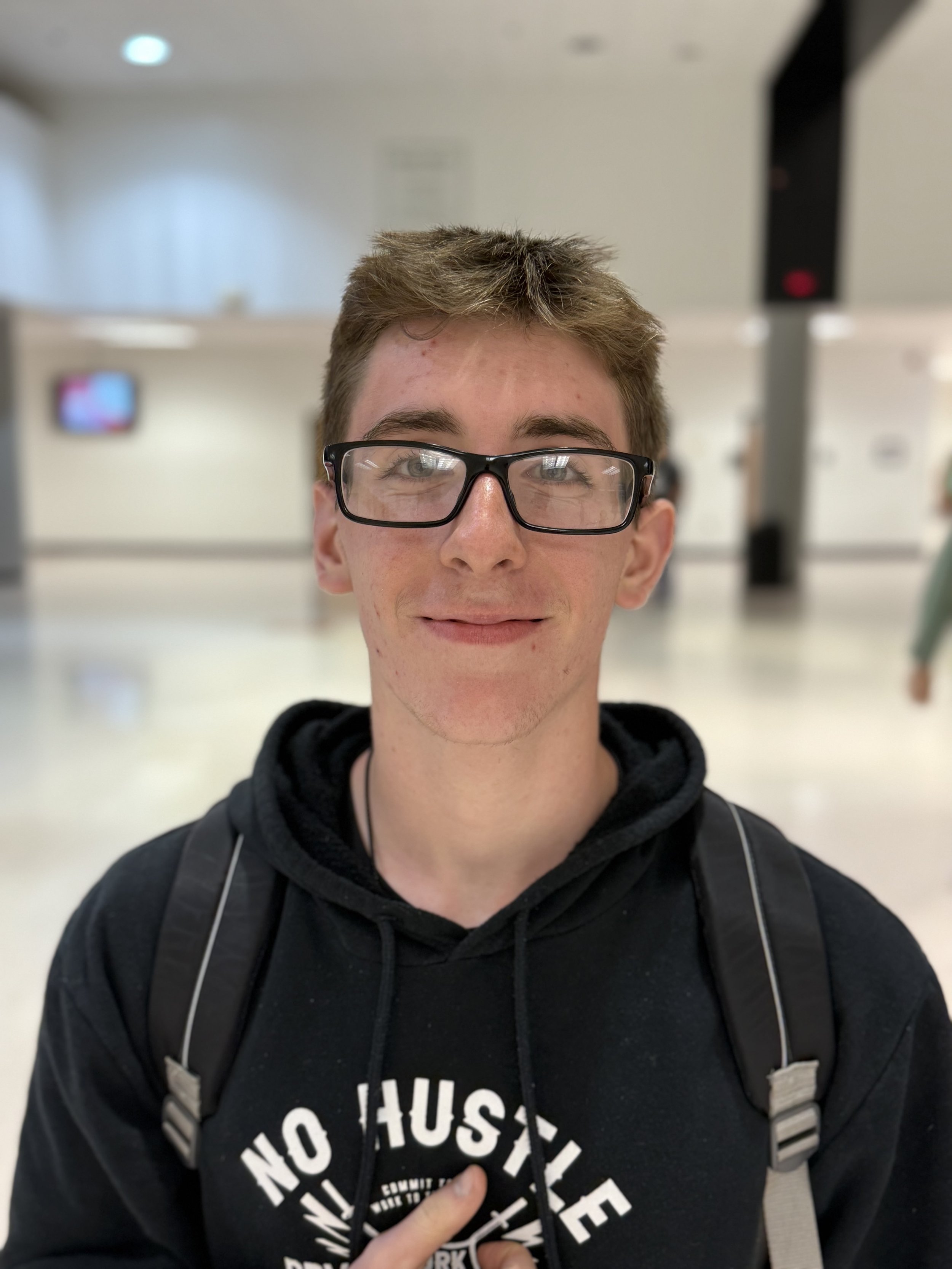 A young man with glasses, short brown hair, wearing a black hoodie with white text, and carrying a backpack, smiling inside a building with a blurred background.