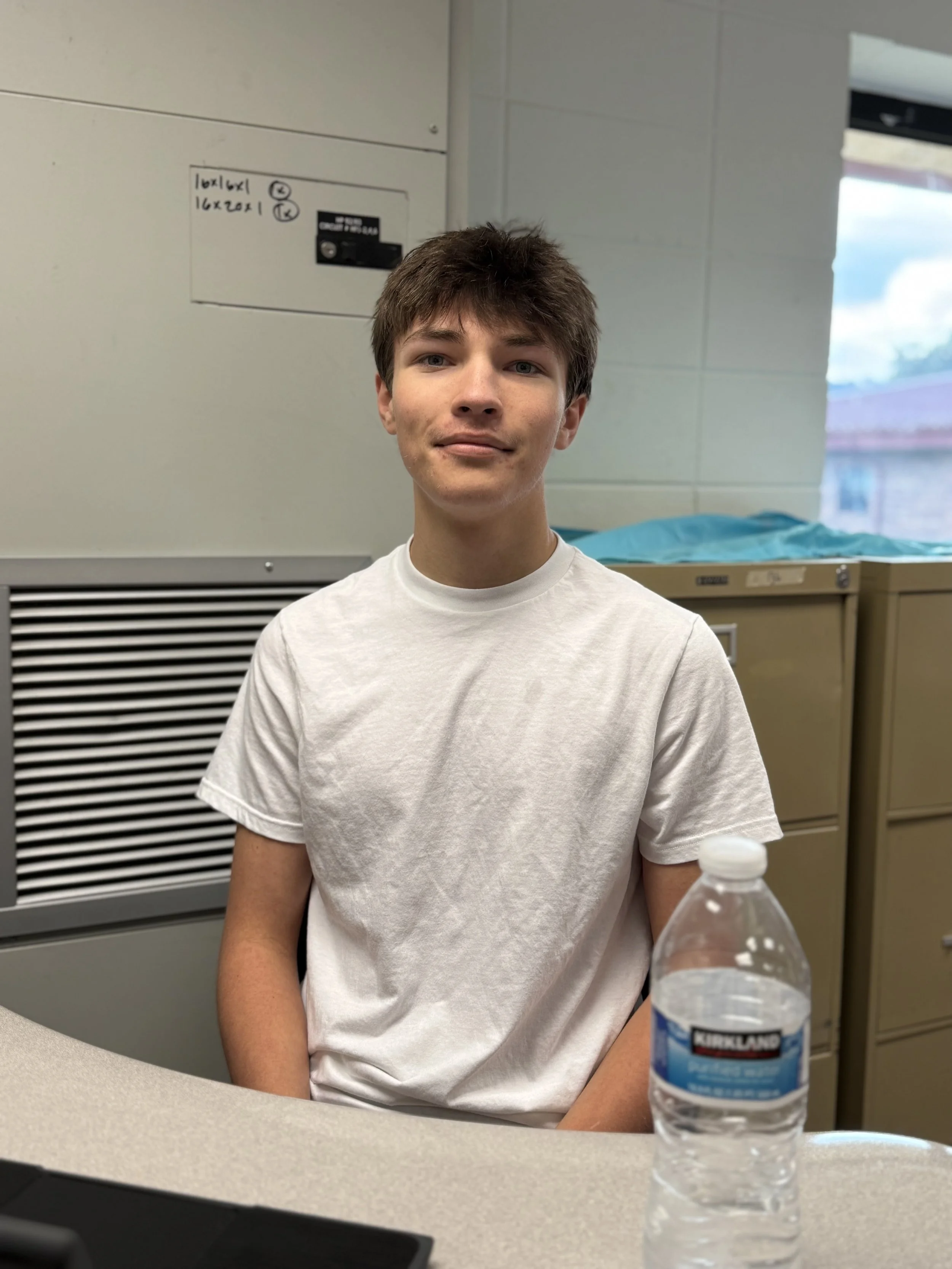 A teenage boy sitting at a desk in a room with beige cabinets and a window. He is wearing a white t-shirt and looking at the camera with a slight smile. There is a water bottle on the desk in front of him.