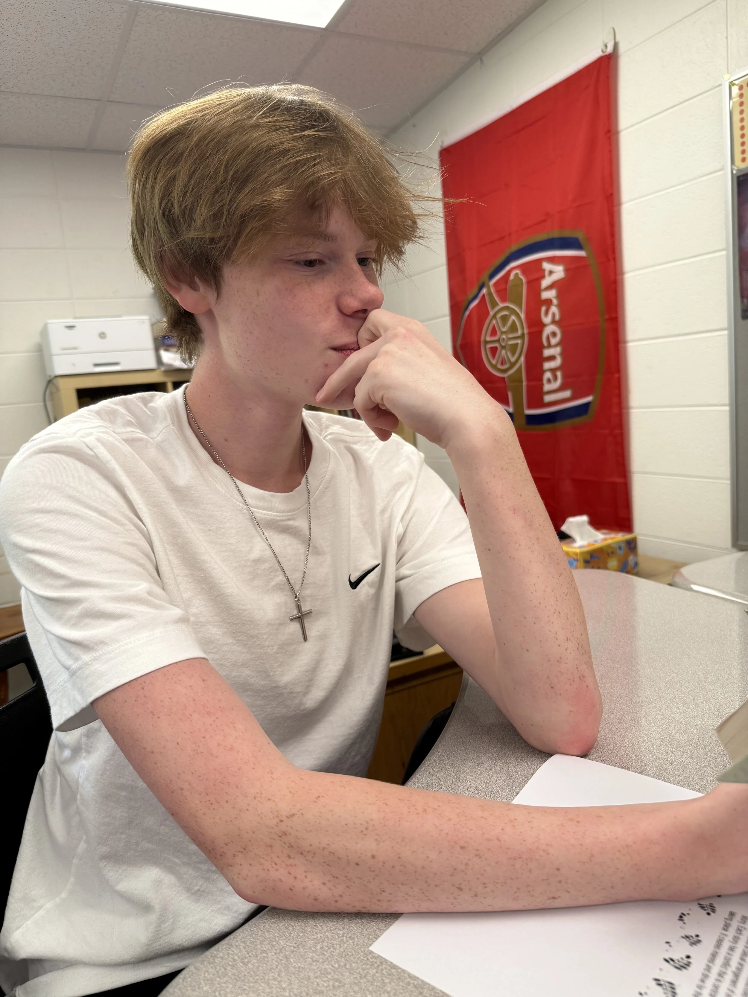 A young person with short red hair and freckles sitting at a desk, smiling, in a classroom or library setting with bookshelves and bags in the background.