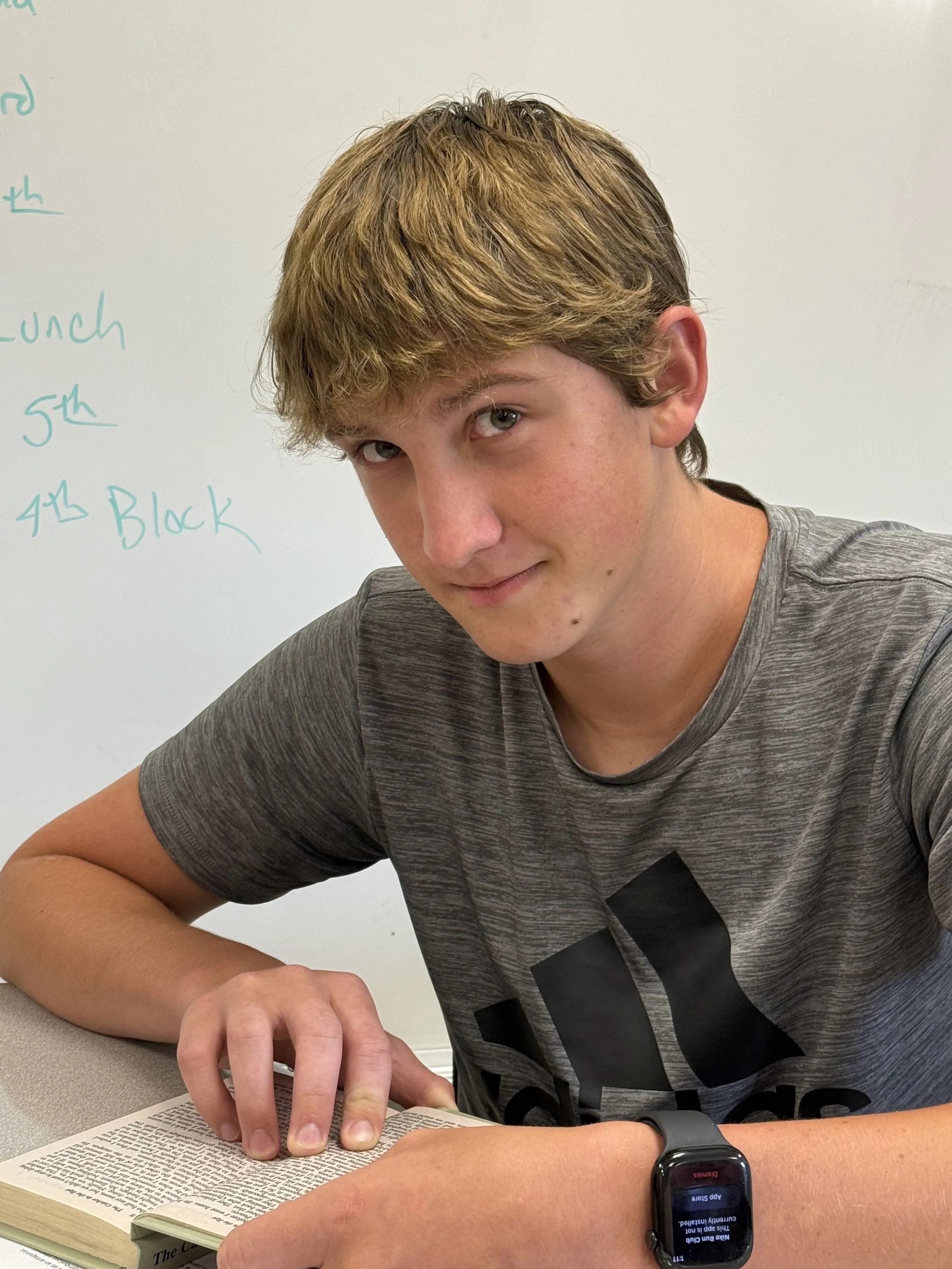 Close-up of a young man with curly blonde hair, wearing a gray hoodie, sitting indoors with a blurred background of people in a classroom or similar setting.