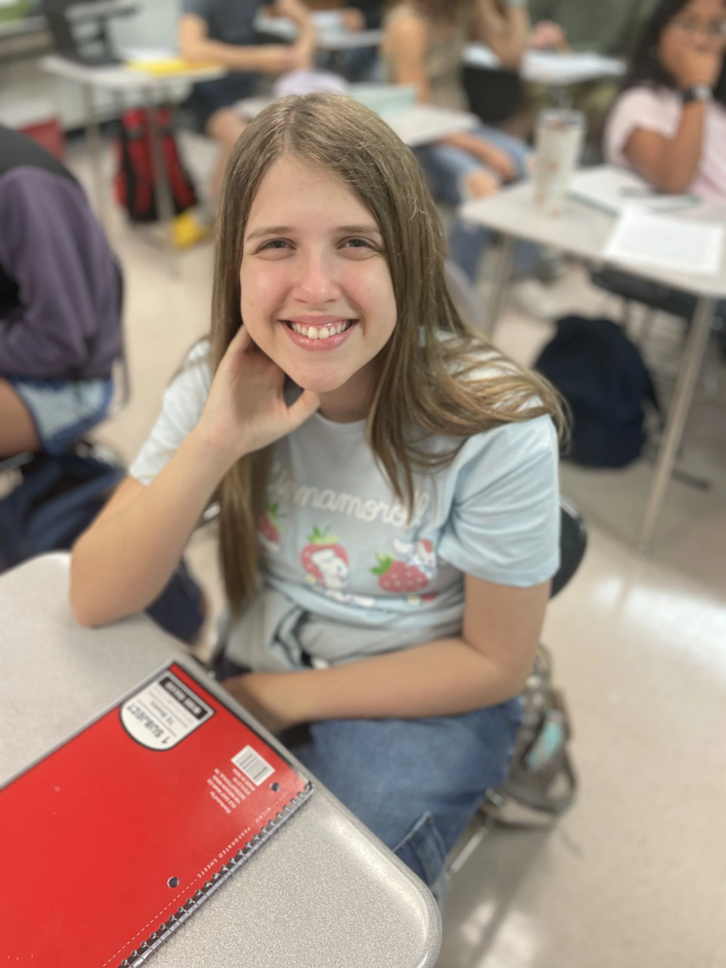 A smiling girl with long light brown hair sitting at a desk in a classroom, wearing a light-colored t-shirt with strawberries and smiling at the camera.