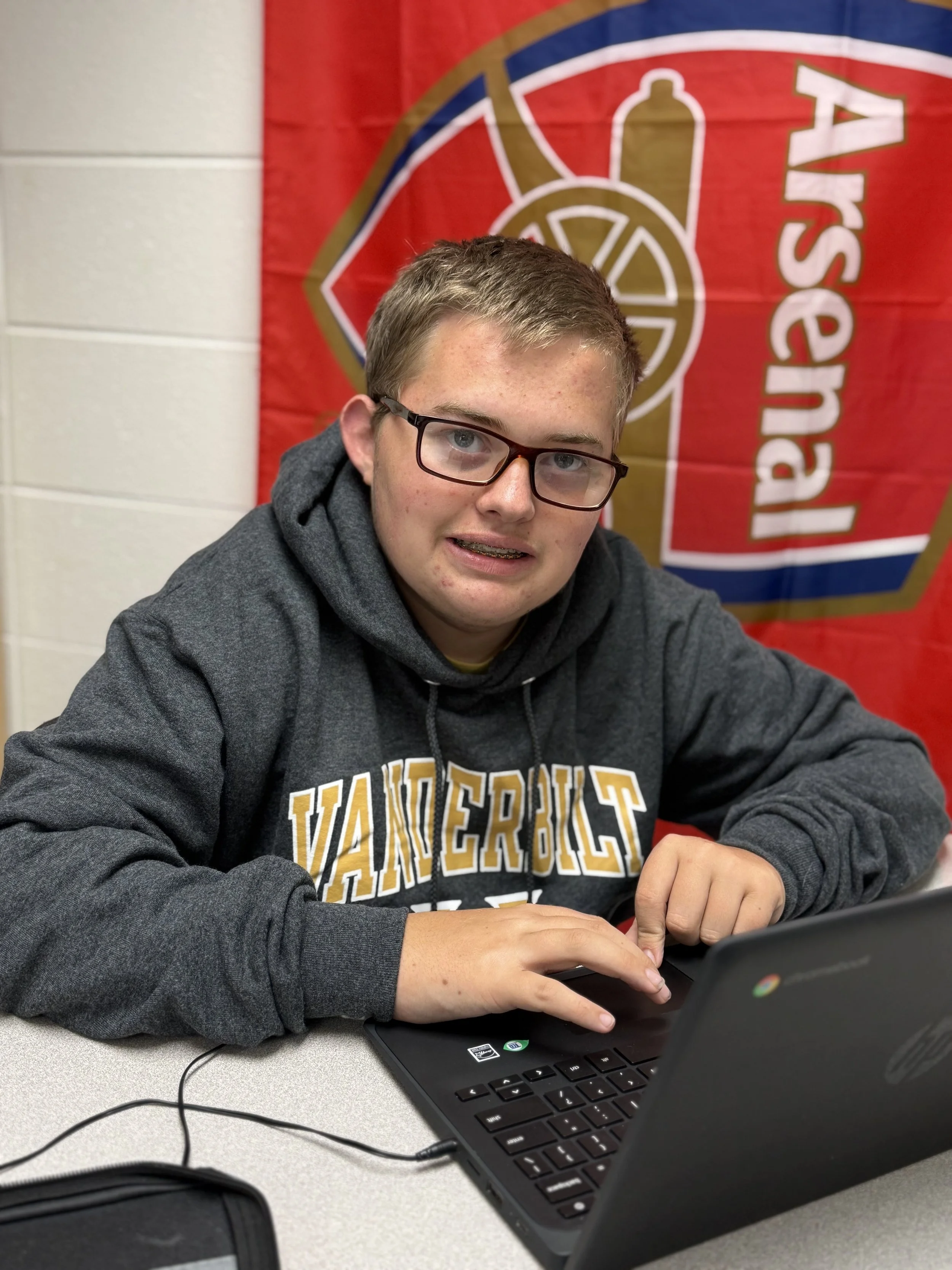A young man with glasses sitting at a table in front of a laptop, wearing a Vanderbilt hoodie, with a red and gold banner behind him that has the Arsenal logo.