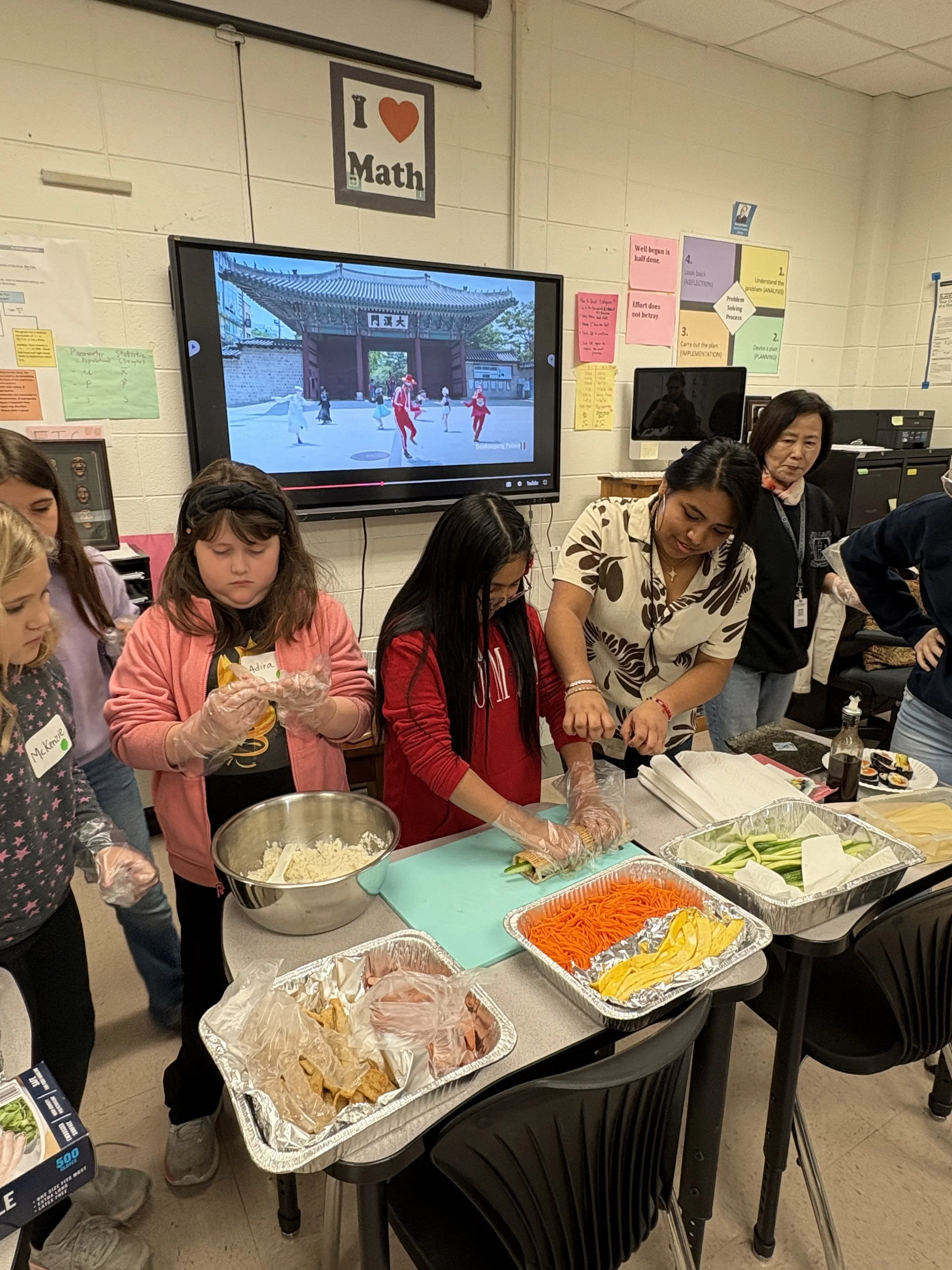 A group of young girls and an adult woman preparing sushi in a classroom with a large screen displaying an Asian temple gate and dancers in the background. The table has trays of sliced vegetables and ingredients for making sushi.