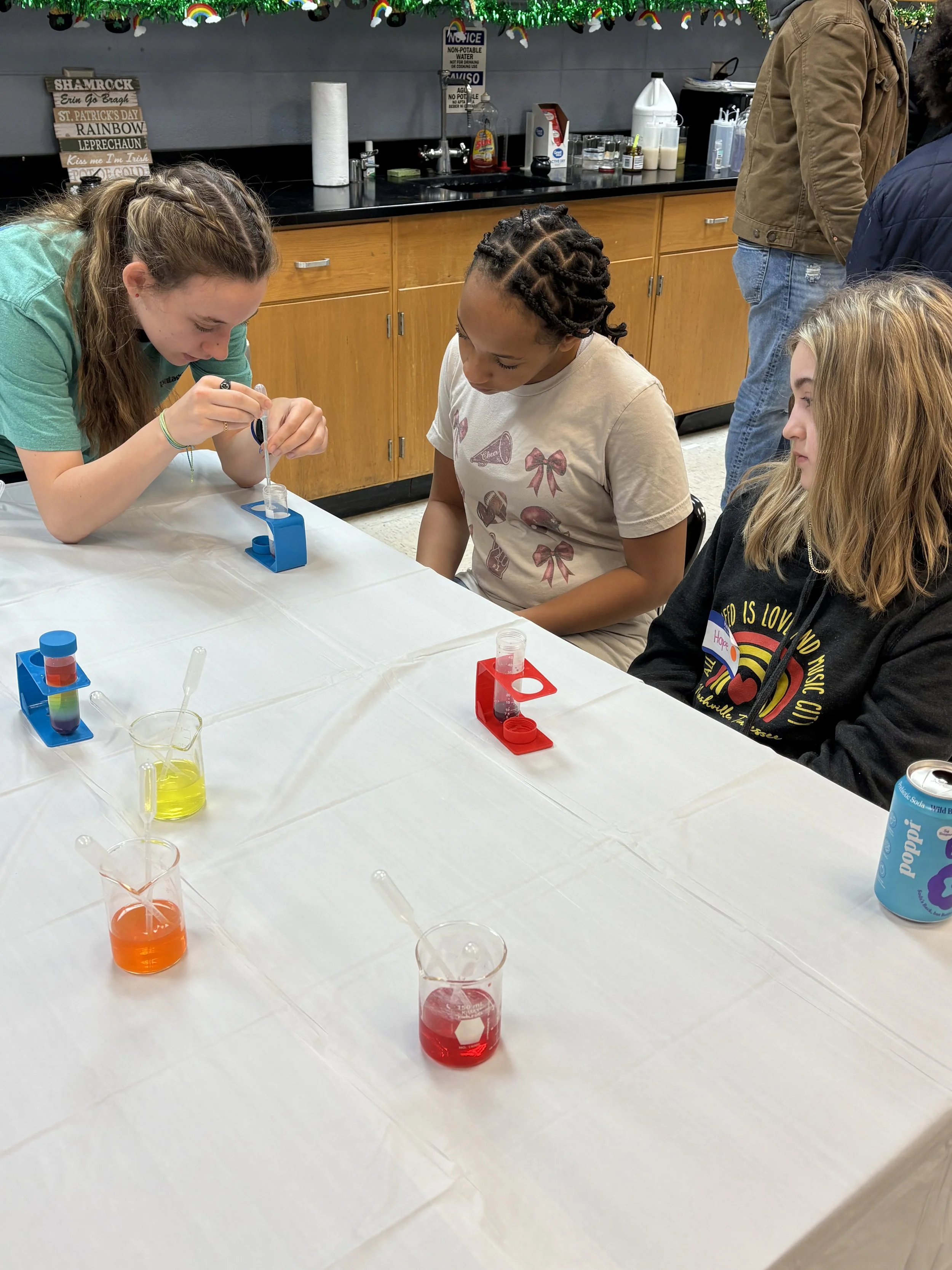 Three girls sit at a table conducting a science experiment with colorful liquids in small beakers and test tubes. One girl is adding a liquid to a test tube, while the other two observe. The table has a white tablecloth and various science supplies. 