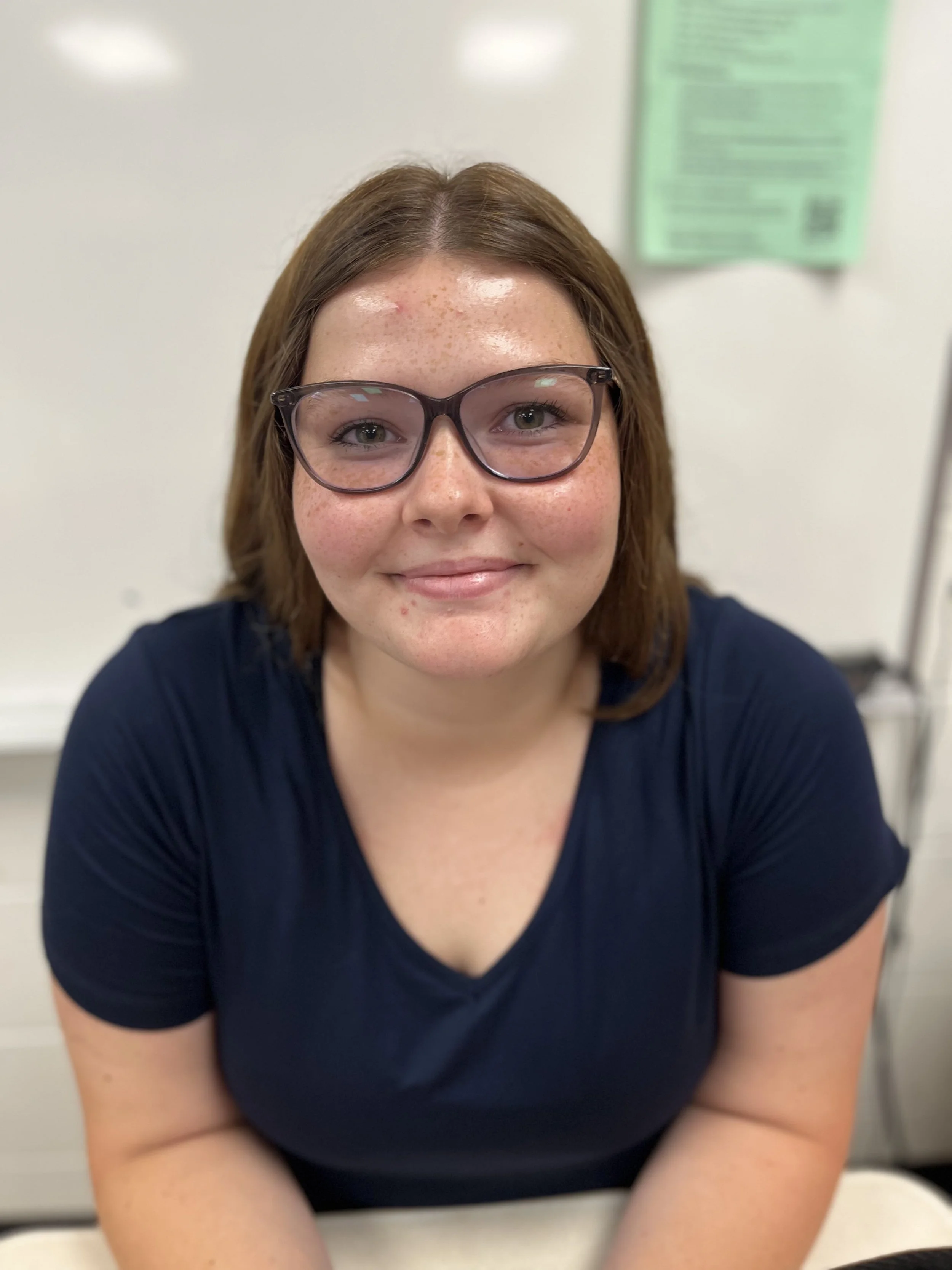 A young woman with glasses and a light purple zip-up jacket sitting at a desk in a classroom, smiling at the camera. In the background, other students are working and talking.