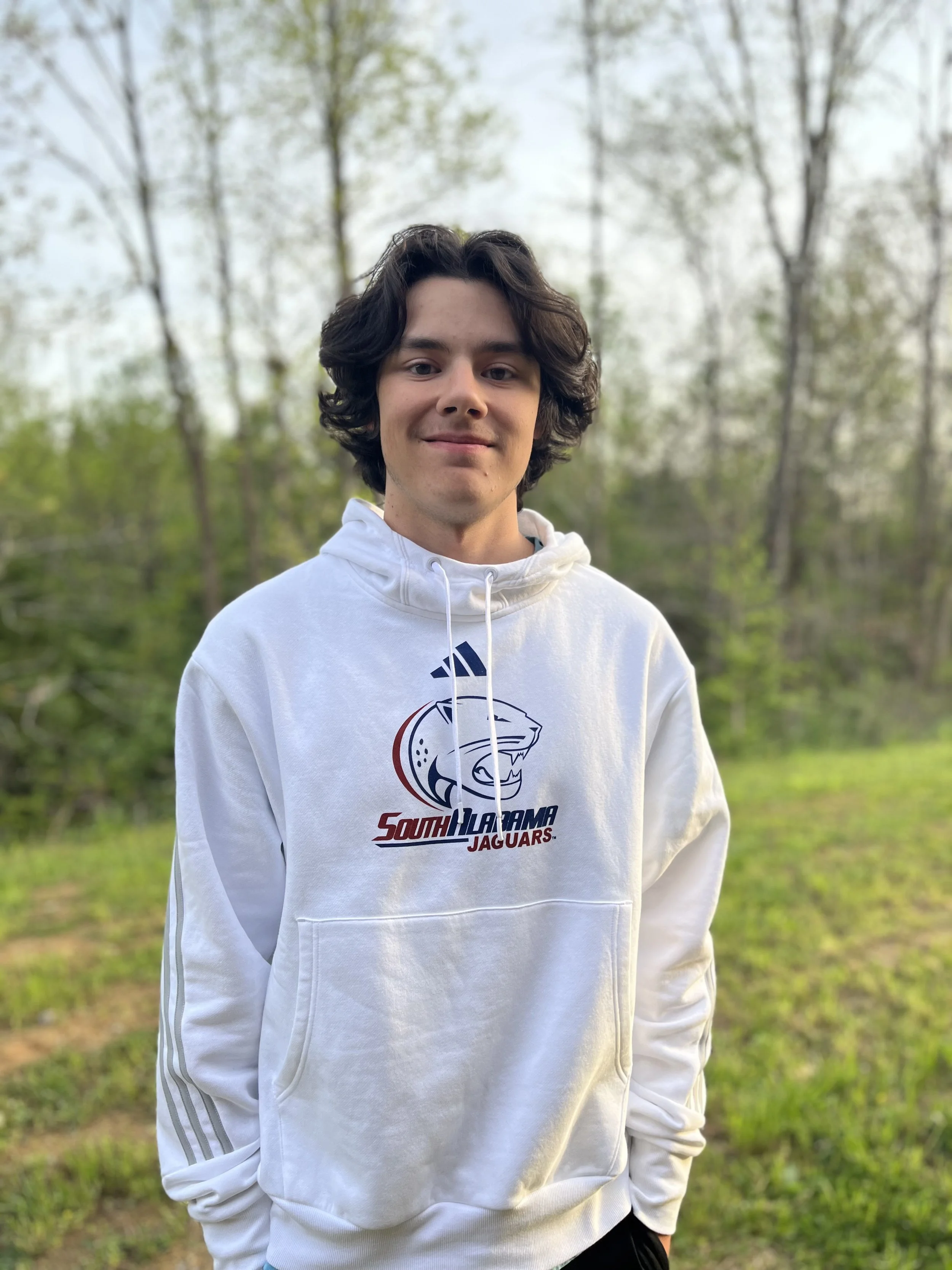Close-up of a young man with shoulder-length dark hair, blue eyes, and light skin, wearing a light green polo shirt, with a blurred background of knives in a knife block.