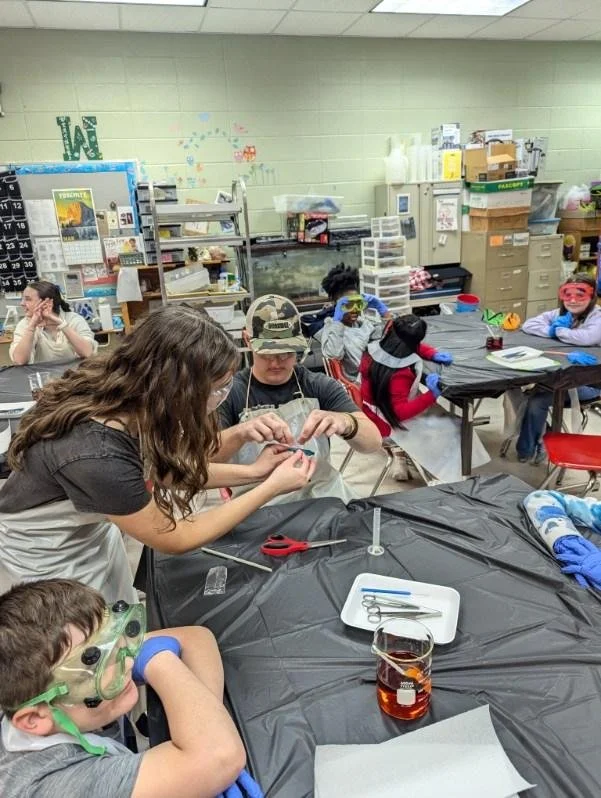 Children and a teacher participating in a science or craft activity in a classroom, wearing safety goggles, with tools and supplies on the table, and classroom backgrounds with storage and decorations.