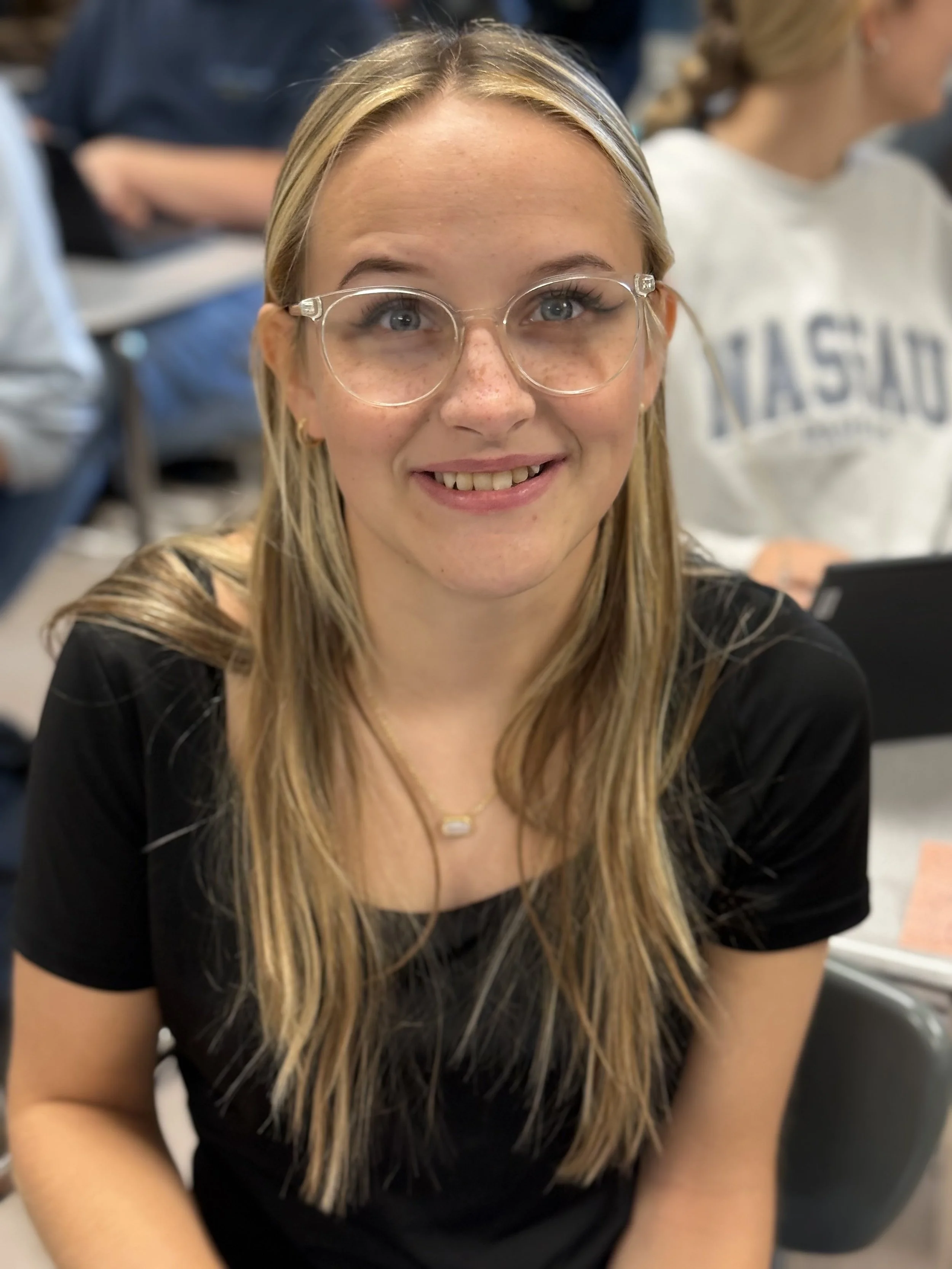 A young woman with long blonde hair, wearing glasses and a black top, smiling at the camera in a classroom or lecture hall setting.