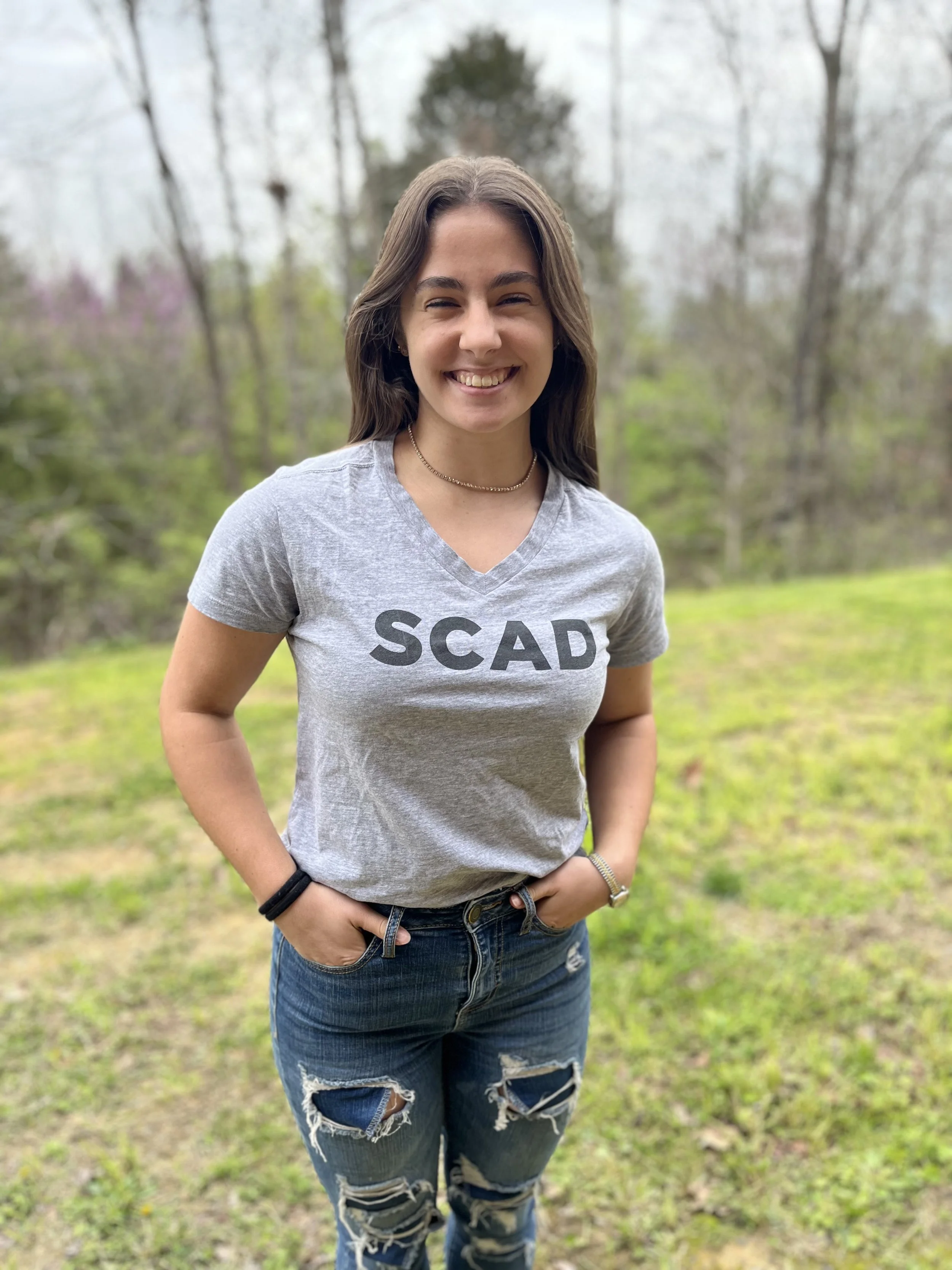 A young woman with long brown hair and a braid, wearing a black sheer top with white polka dots, sitting in a classroom setting.