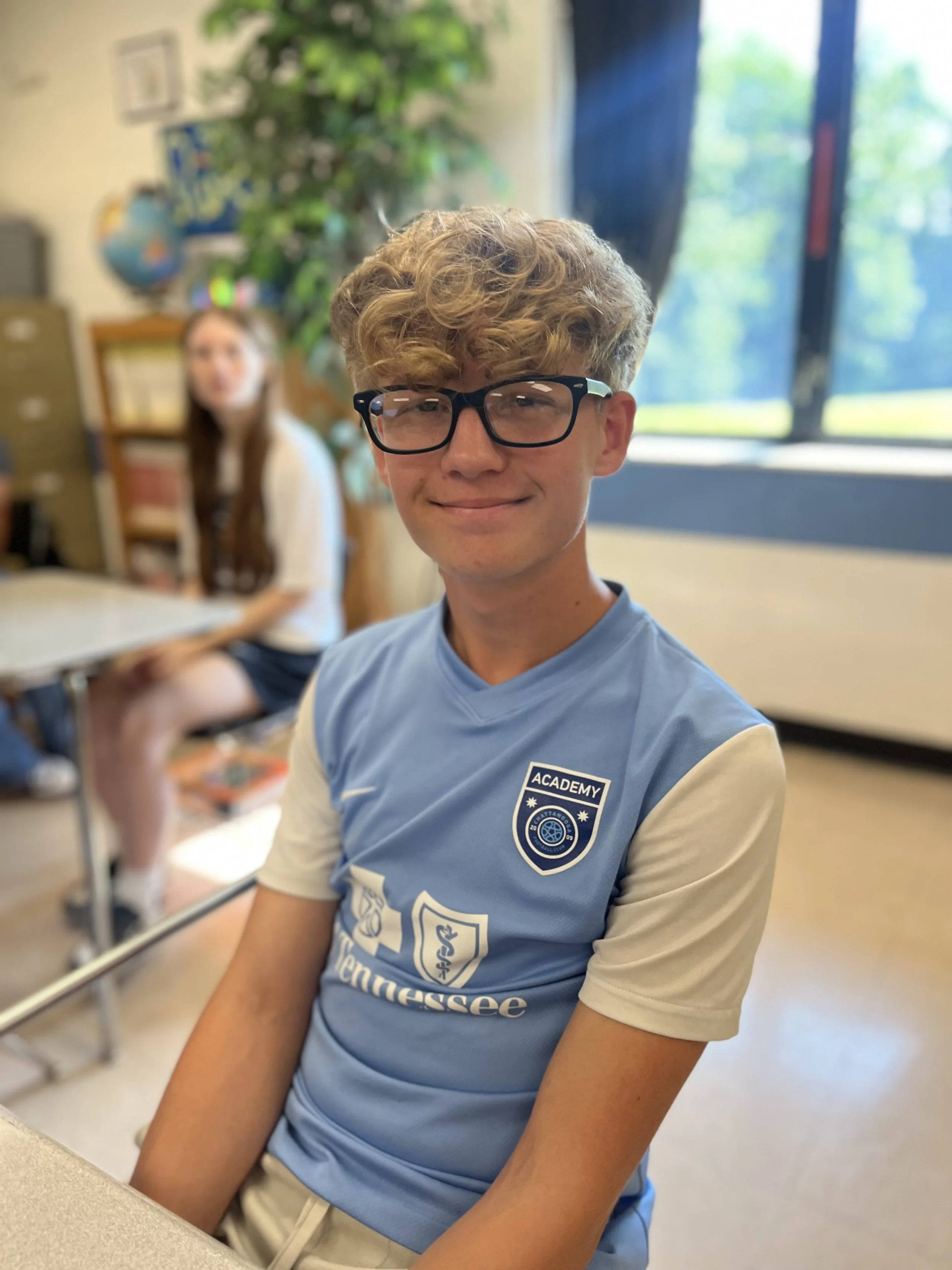 A young boy with red glasses and light brown hair wearing a blue and red jacket sits at a desk in a classroom, with other students in the background.