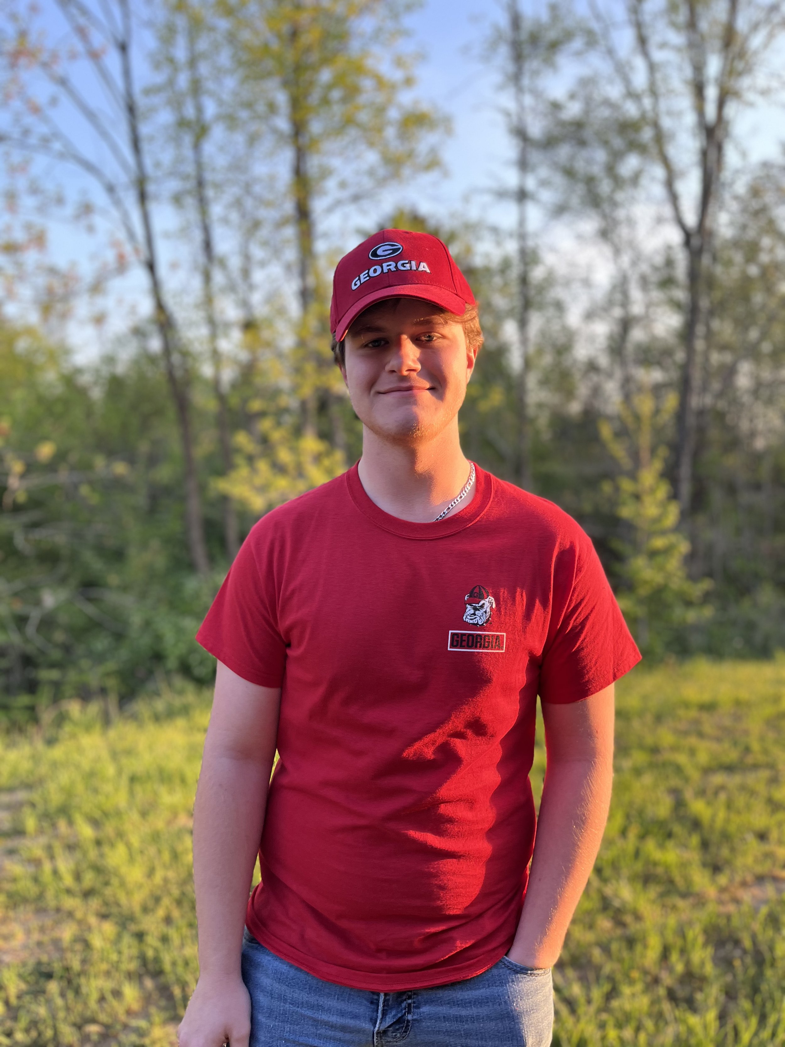 Young man with short light brown hair smiling in a classroom or library setting with blurred background showing other students, bookshelves, and a blue backpack.