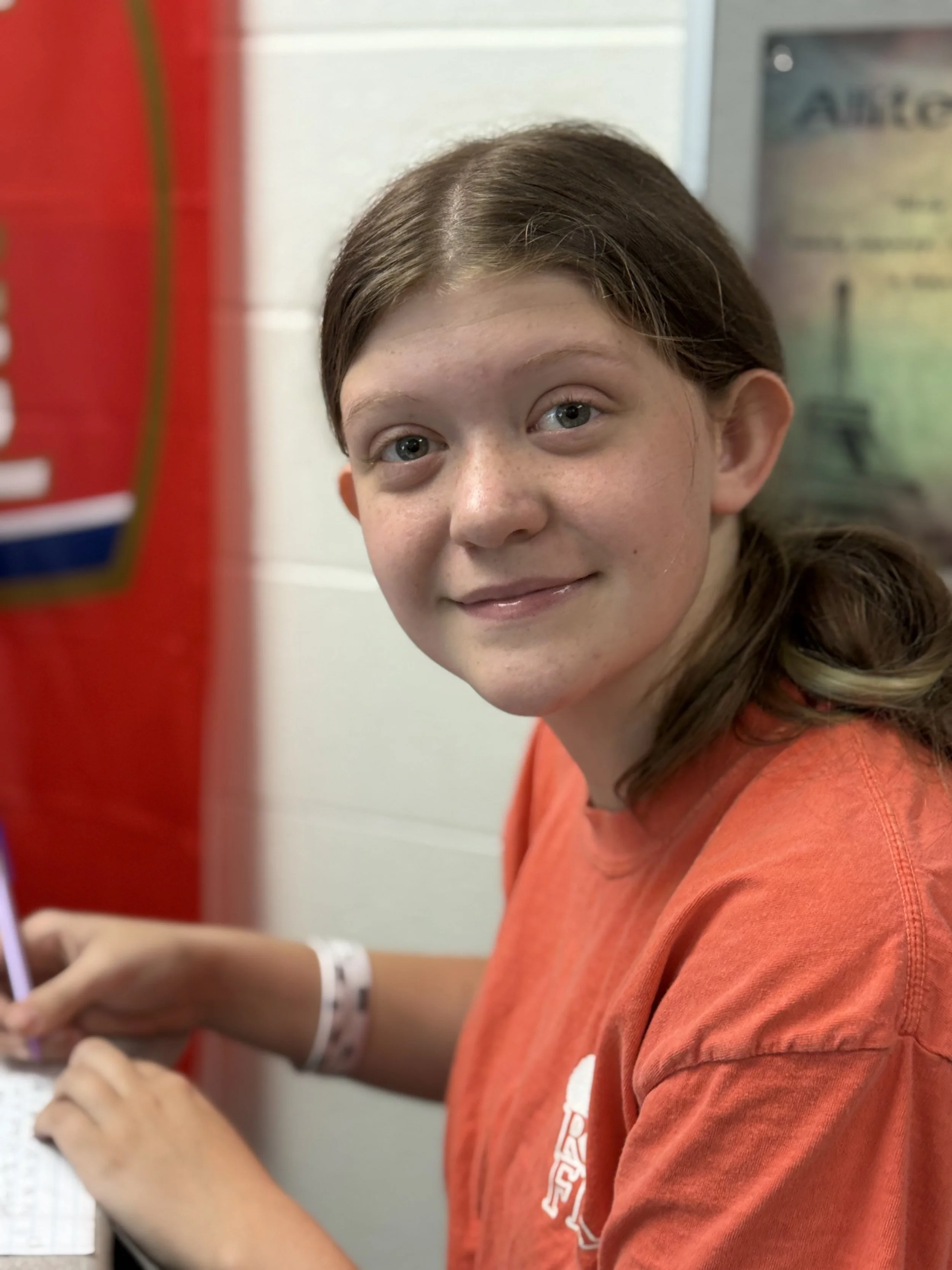 A young girl with brown hair and green eyes smiling at the camera, wearing an orange shirt, and sitting at a desk with a laptop.