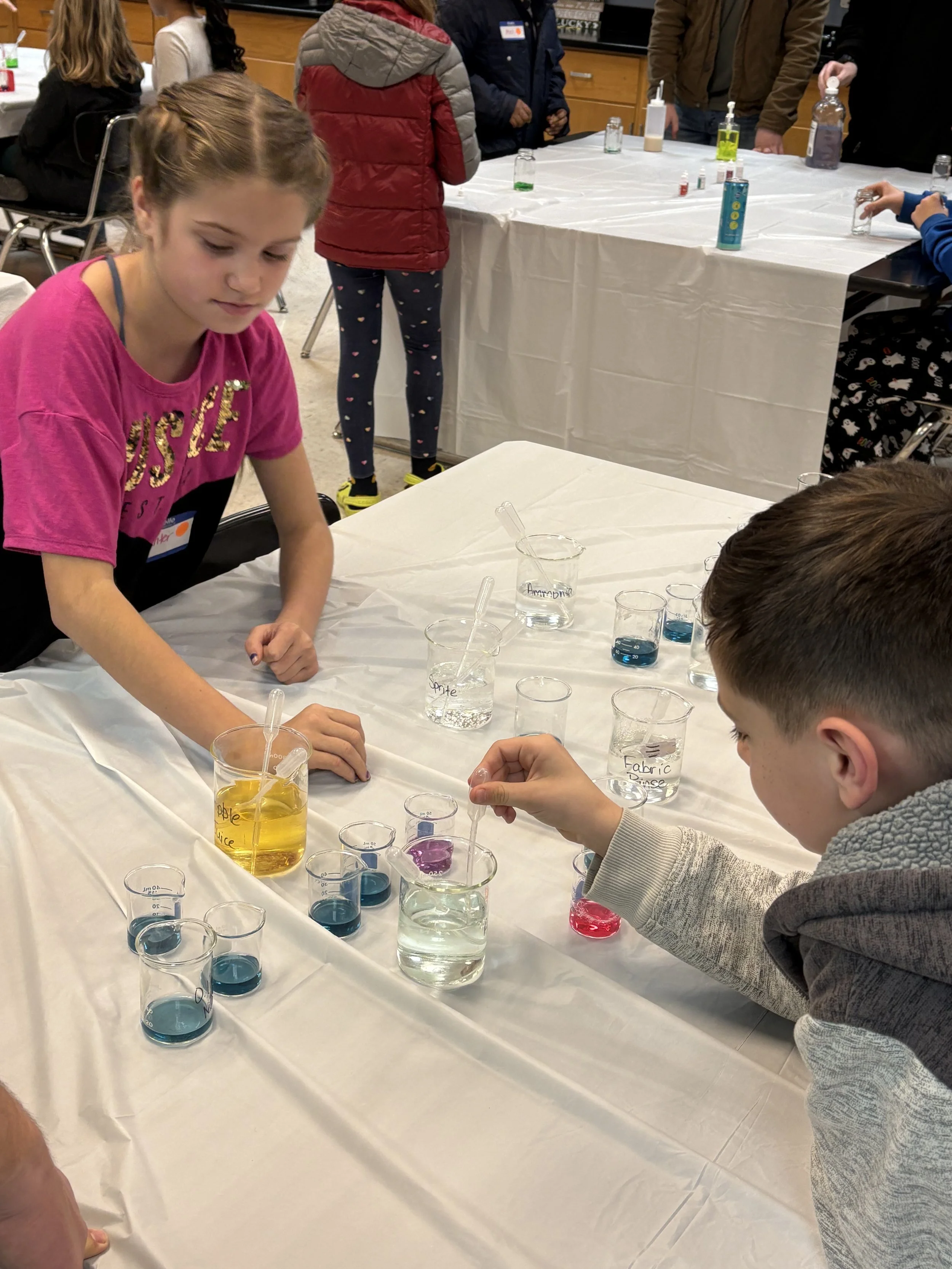Children conducting a science experiment with colorful liquids in beakers on a table covered with a white cloth.