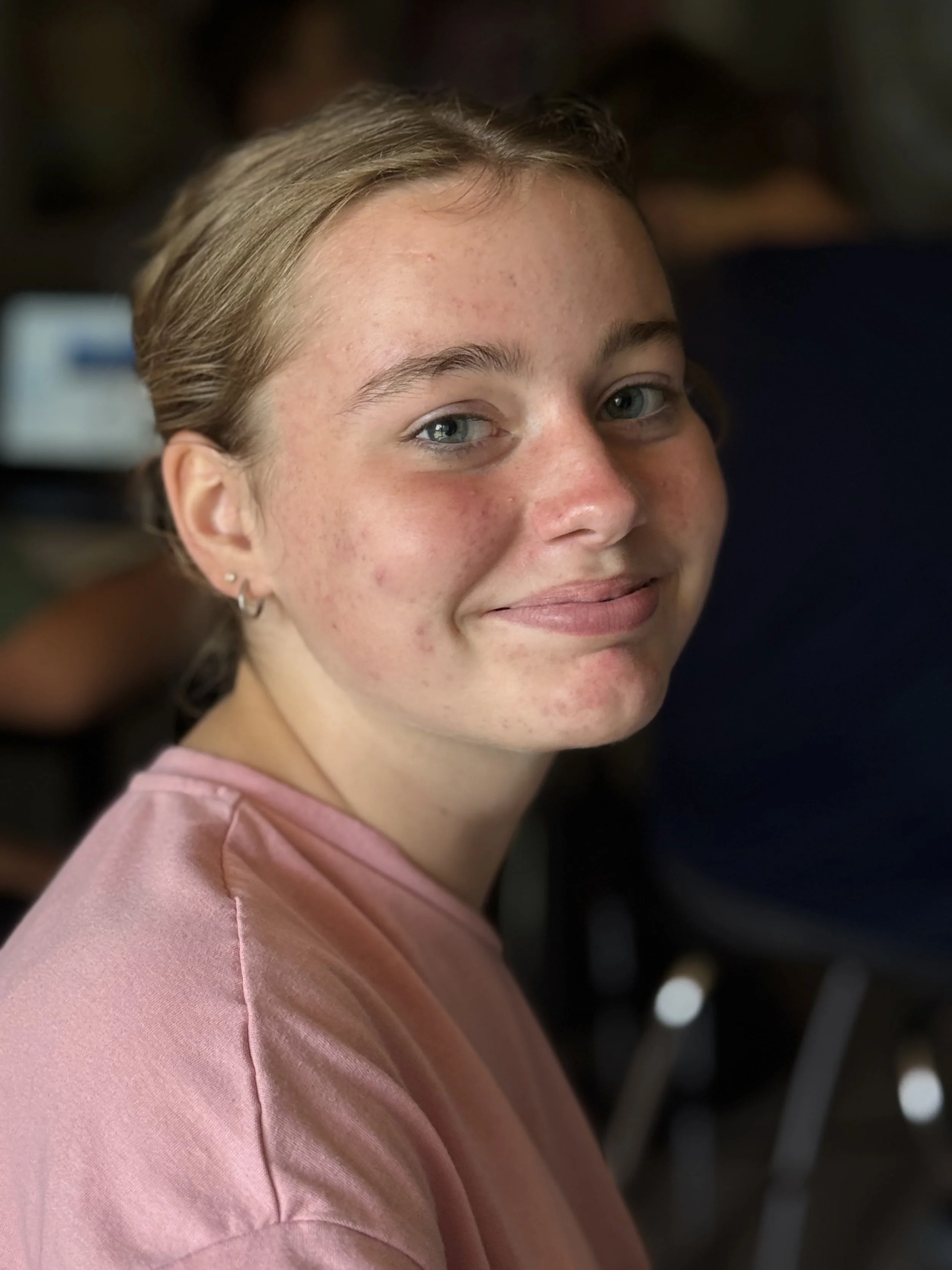 Close-up of a young woman with blonde hair, blue eyes, and a slight smile, wearing a pink shirt.