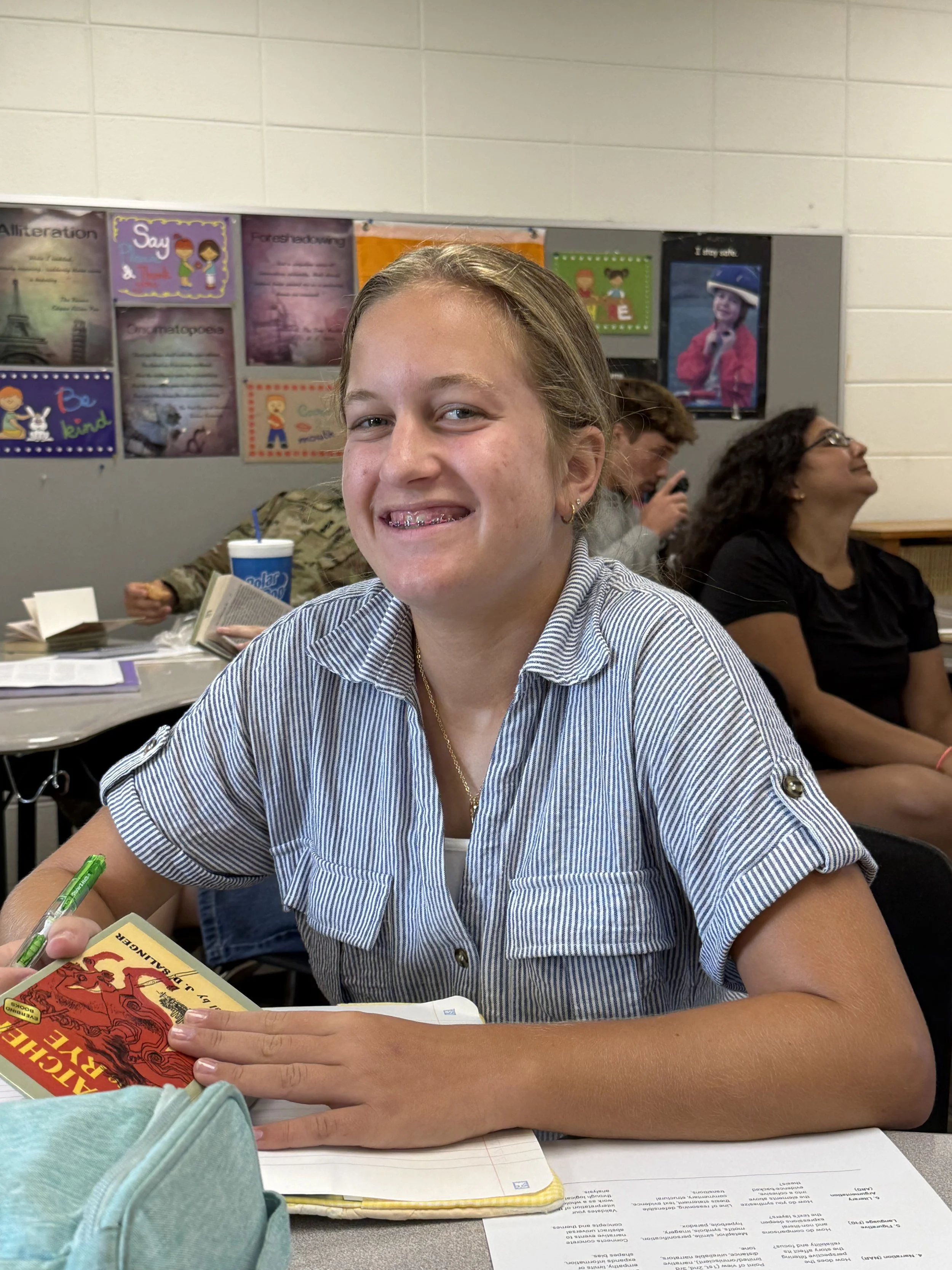 A smiling girl sitting at a table in a classroom, wearing a black hoodie with white text, with school supplies and other students visible in the background.