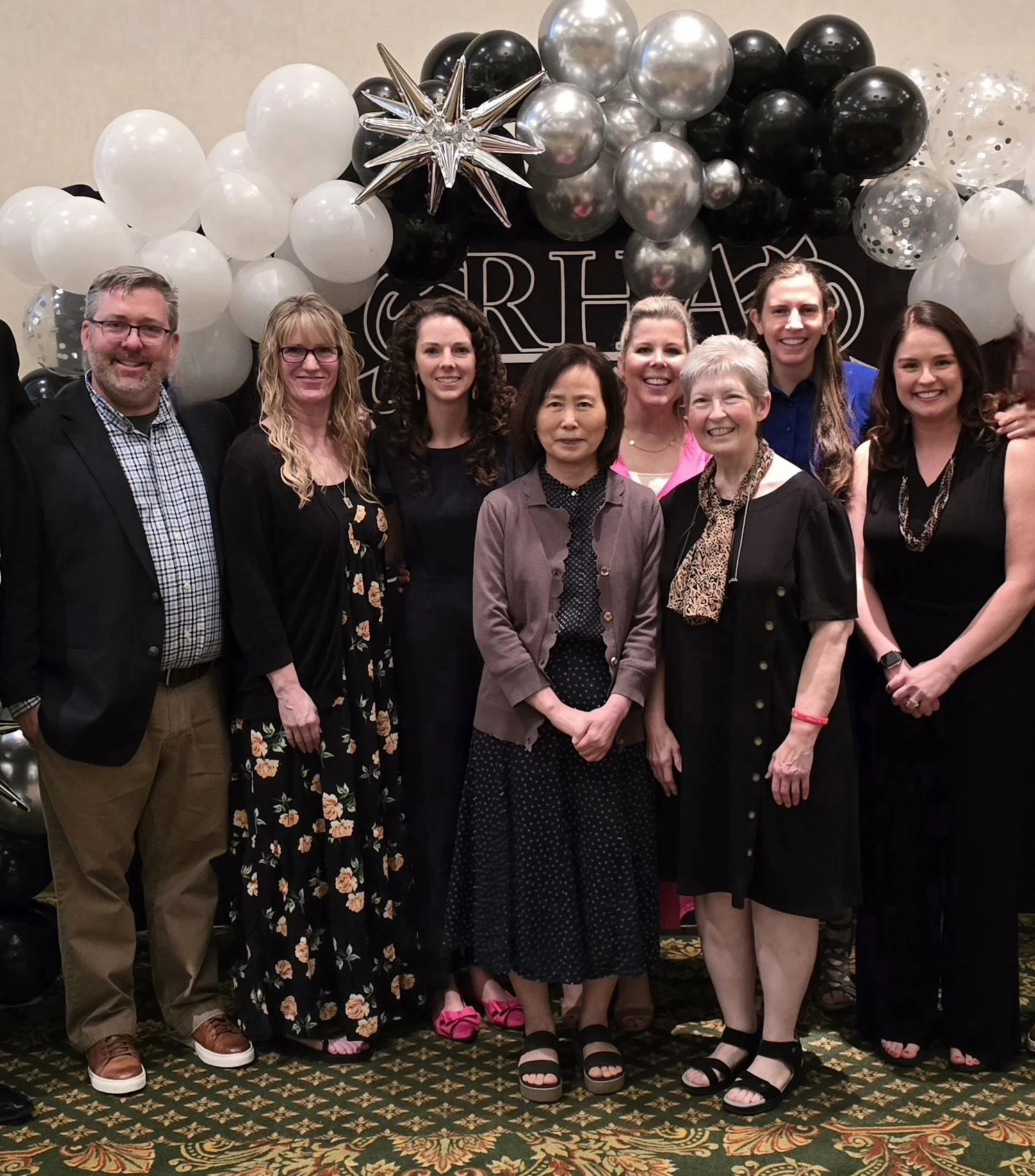 Group of eight people, six women and two men, standing in front of a festive balloon decoration with black, white, silver, and clear balloons, a silver star-shaped balloon, and a black backdrop with white lettering.