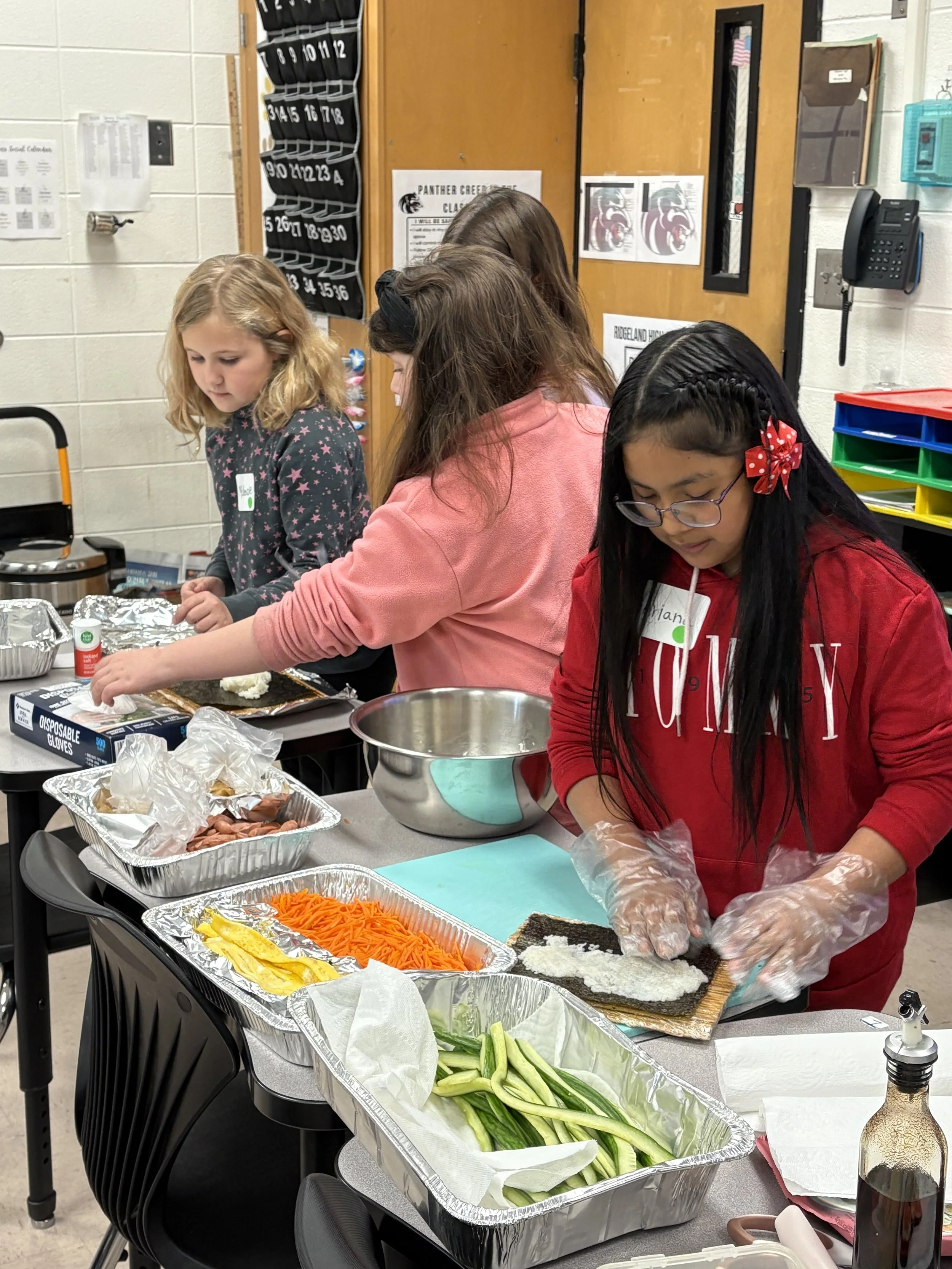 Four girls preparing sushi at a classroom table with trays of vegetables, rice, and seaweed.