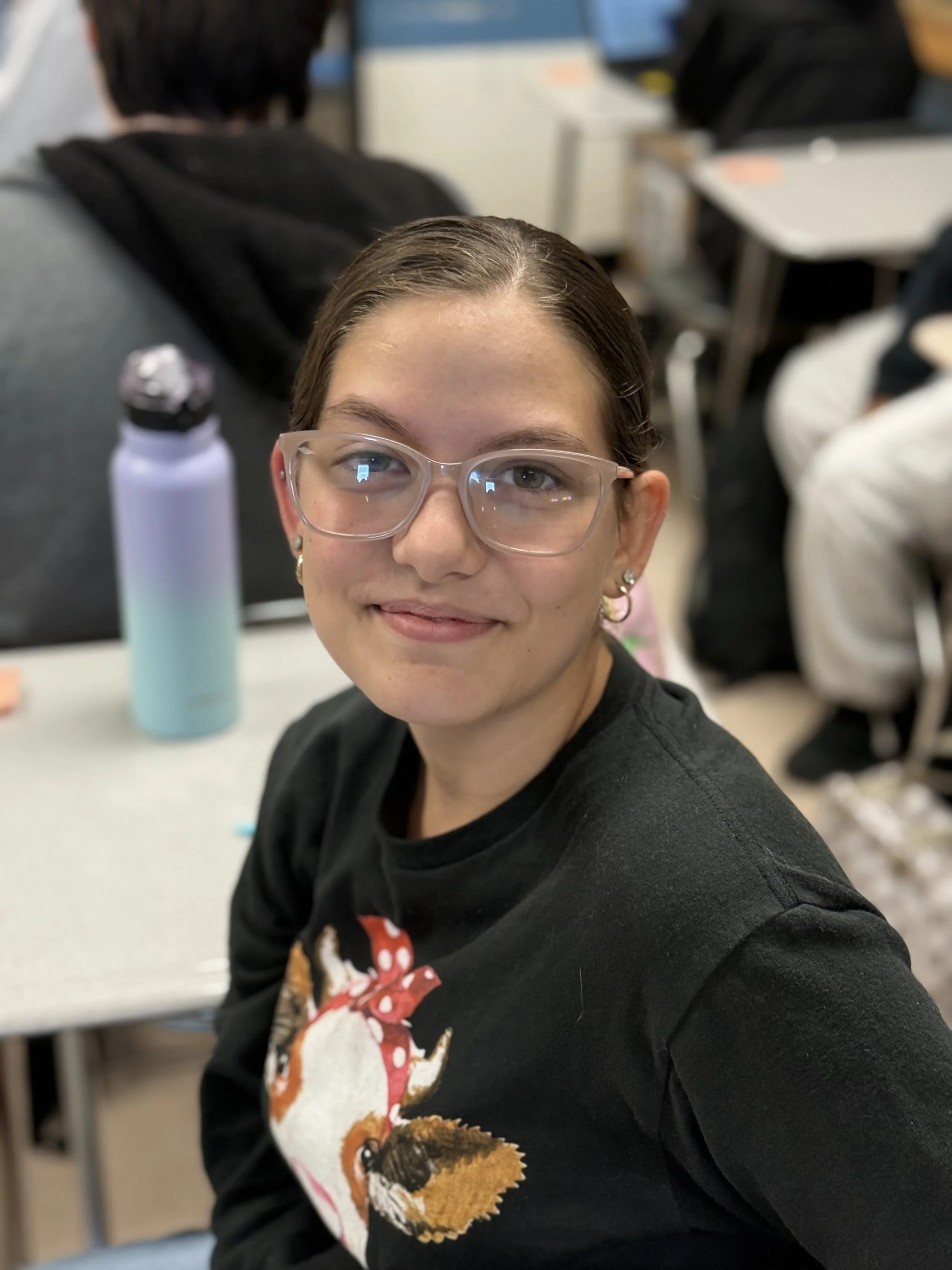 A young woman with glasses and earrings smiling at the camera in a classroom setting, with desks and other students in the background.'