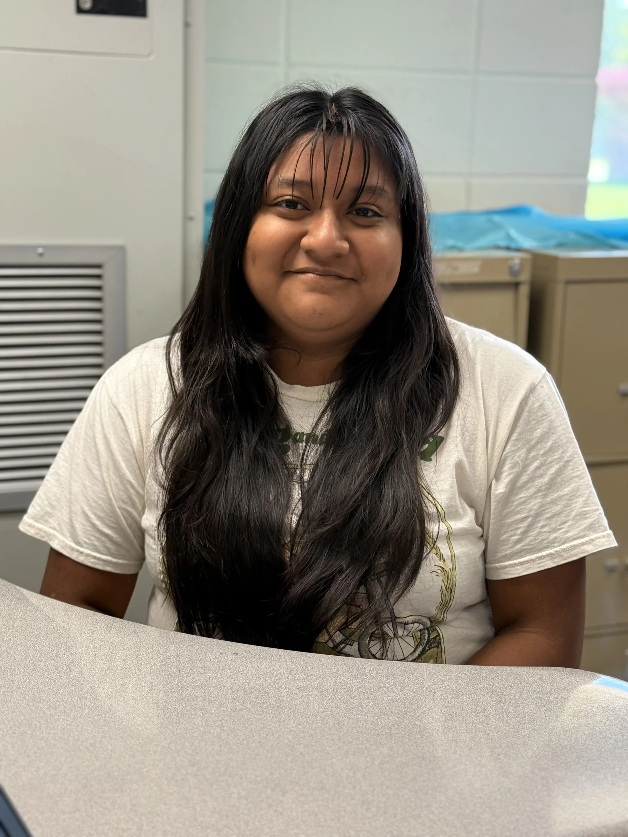 A woman with long dark hair and a white T-shirt sitting at a table in a room with a gray wall and cabinets in the background.