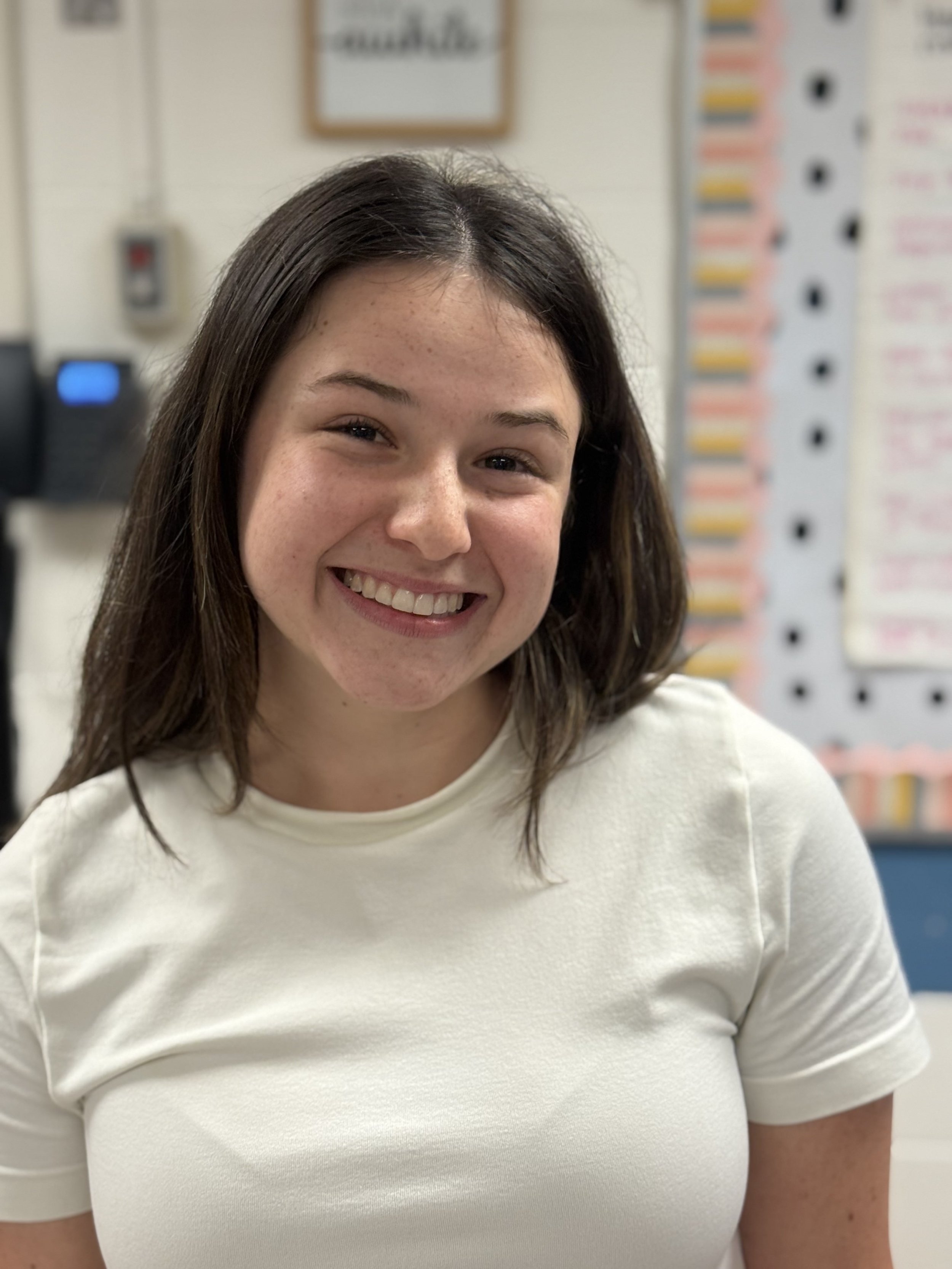 Smiling young woman with shoulder-length brown hair wearing a white t-shirt in an indoor setting with a background of bulletin boards and office equipment.
