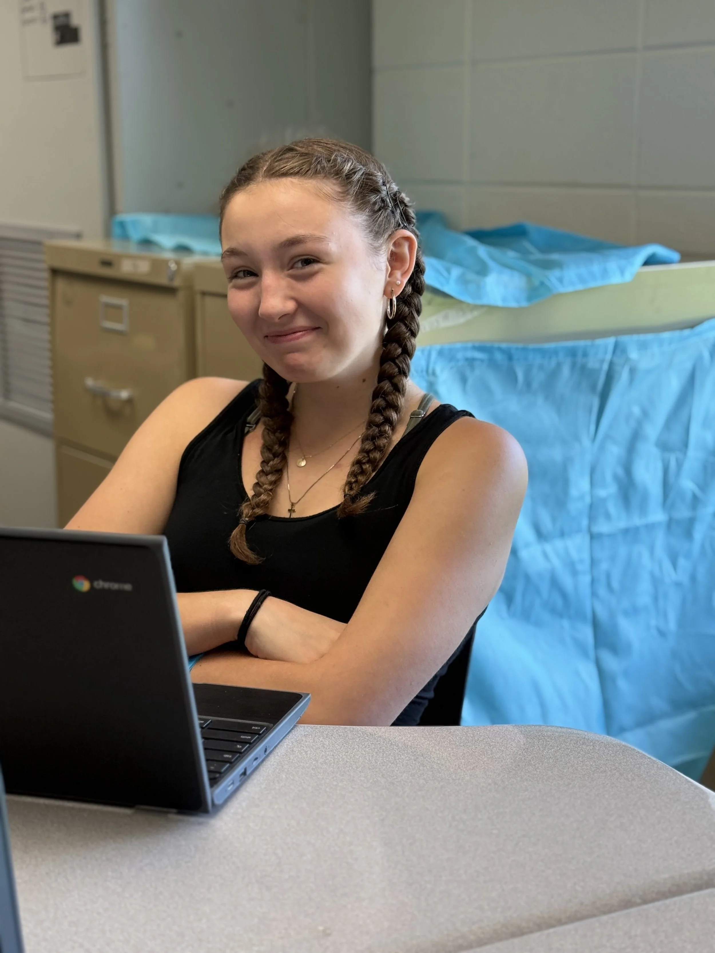A young woman with braided hair sitting at a table with a laptop in front of her, smiling. She wears a black sleeveless top and earrings, with a backpack and medical curtains or sheets in the background.