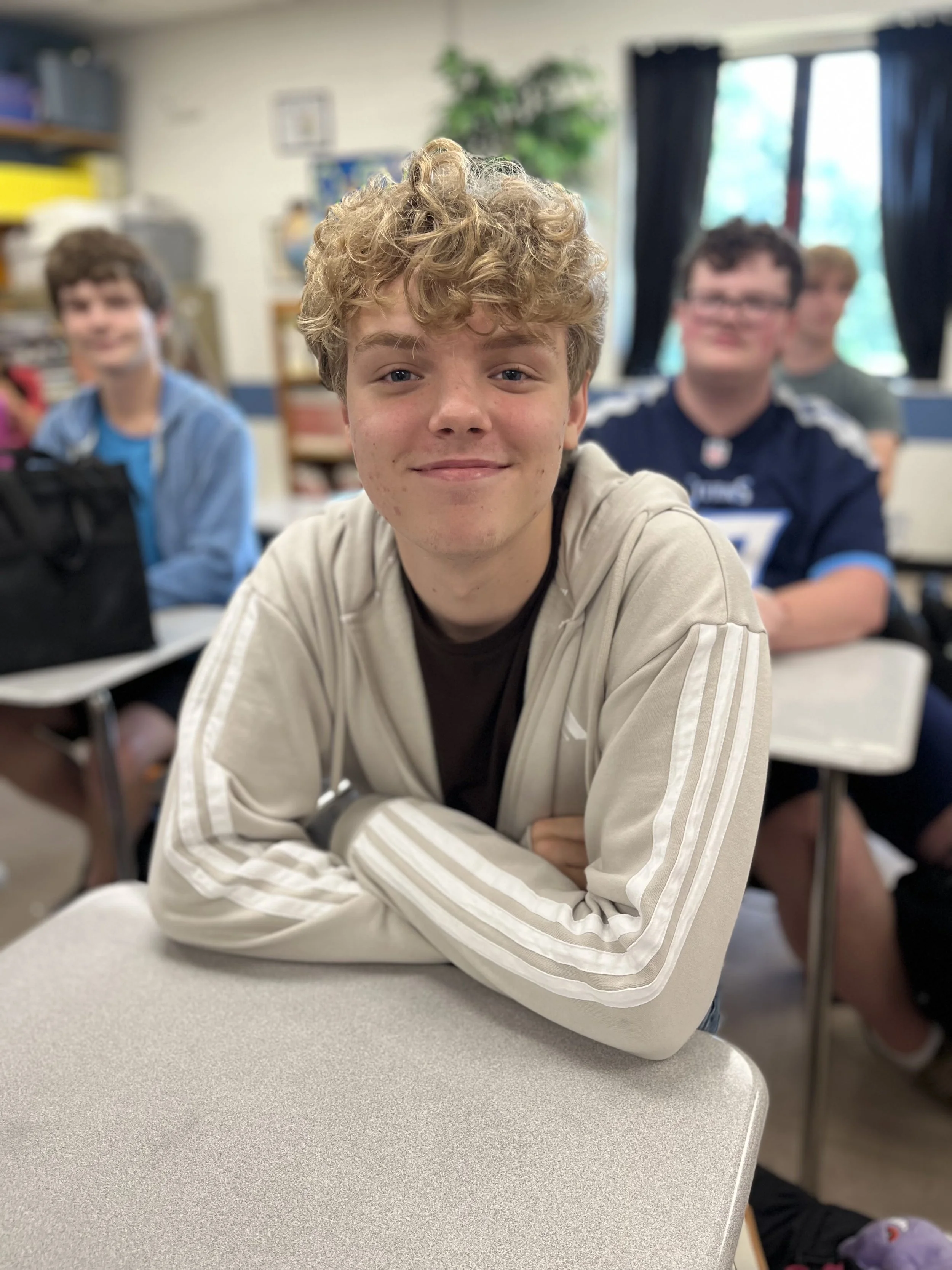 A young person with curly blonde hair and fair skin, wearing a black Nike jacket, smiling softly in an indoor setting.