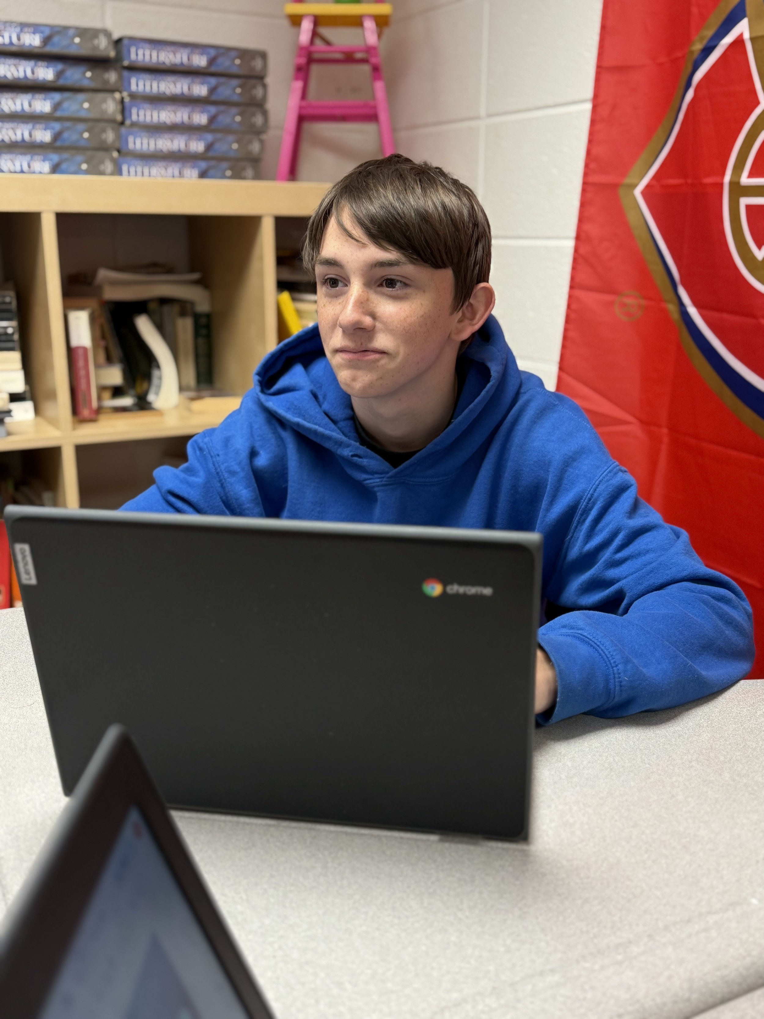 A teenage boy with brown hair and freckles sitting at a table in a classroom, wearing a blue hoodie, working on a Chromebook laptop.