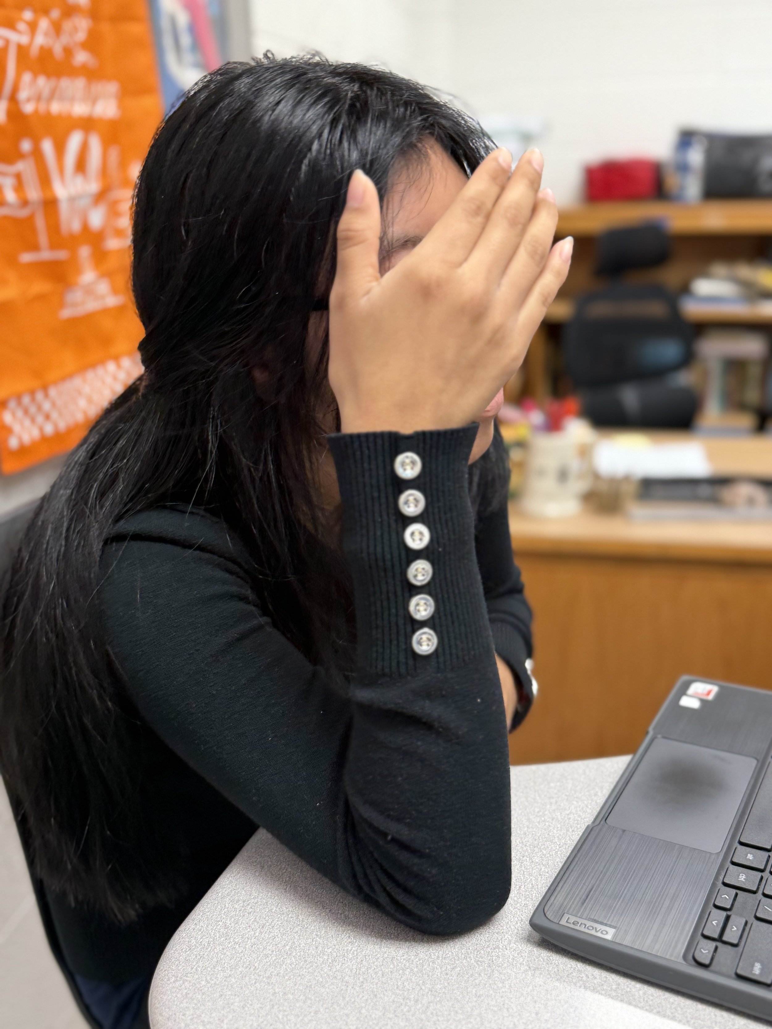 Person with long black hair sitting at a desk covering her face with her right hand, in front of a laptop, in an office or classroom setting.