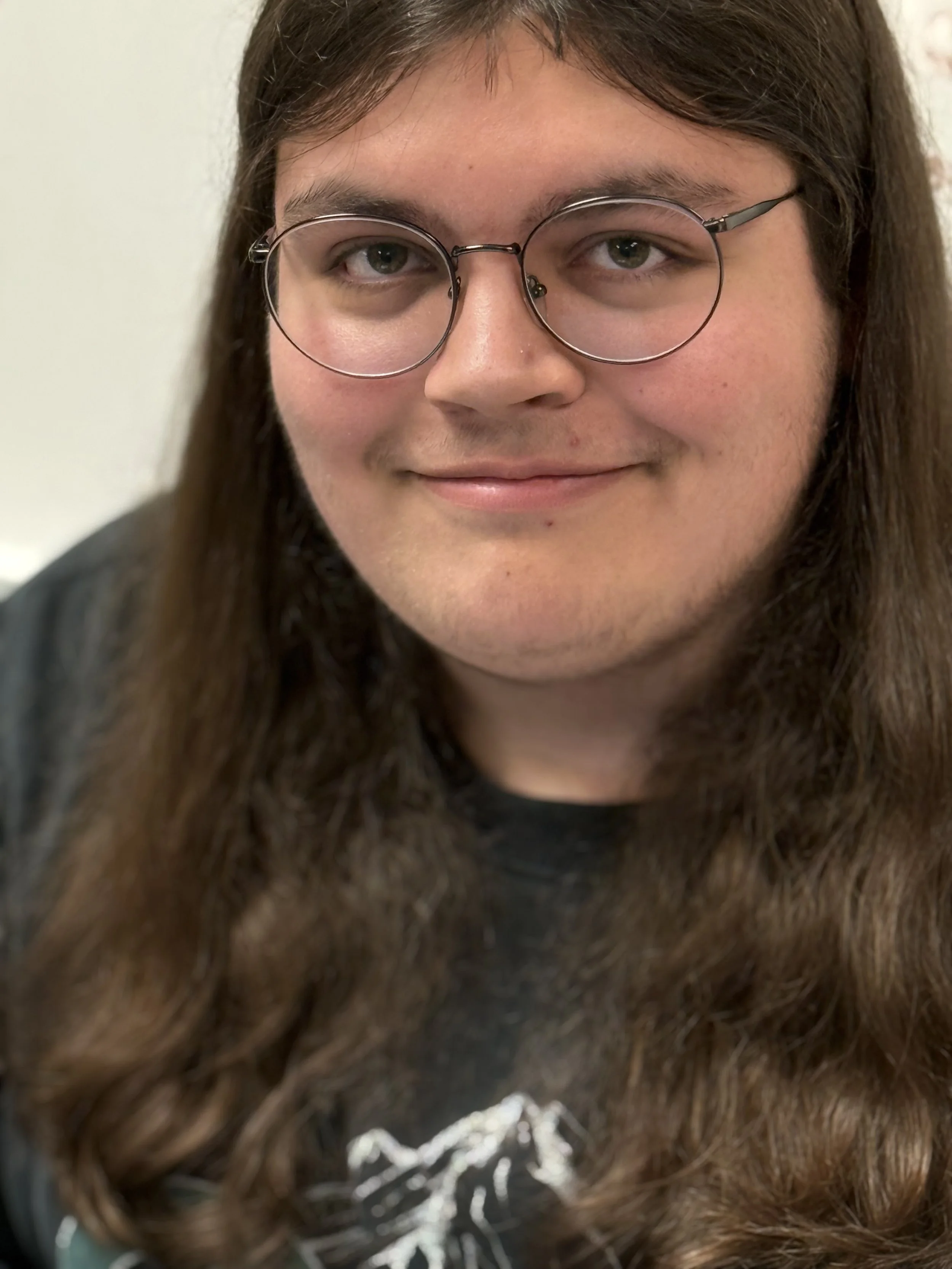 Close-up of a young person with long brown hair, wearing glasses, smiling, with a black t-shirt featuring a white mountain graphic.
