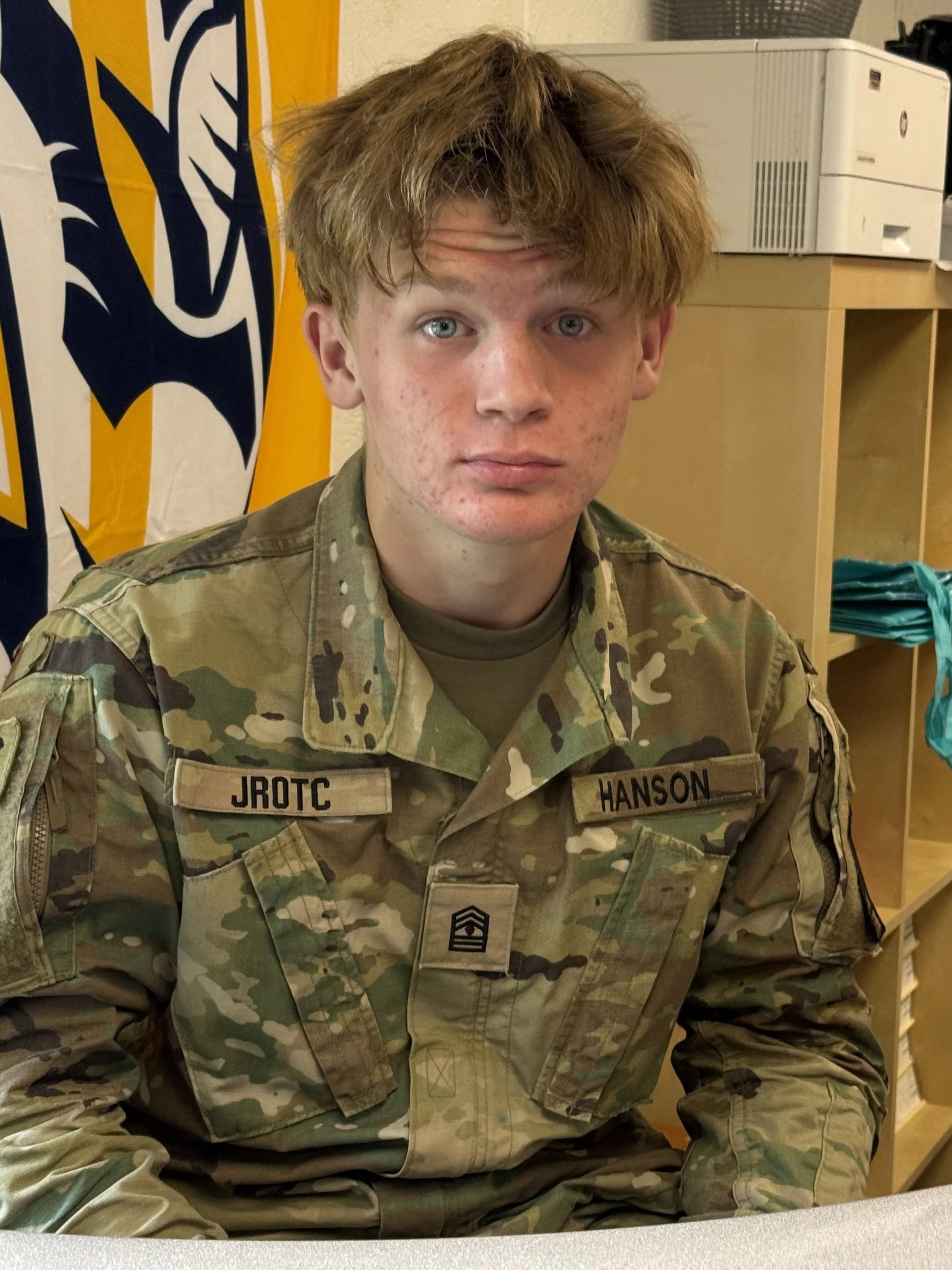 A teenage boy with red hair and blue eyes, wearing a tan t-shirt, leaning over a laptop on a desk, looking at the camera with a surprised or curious expression.