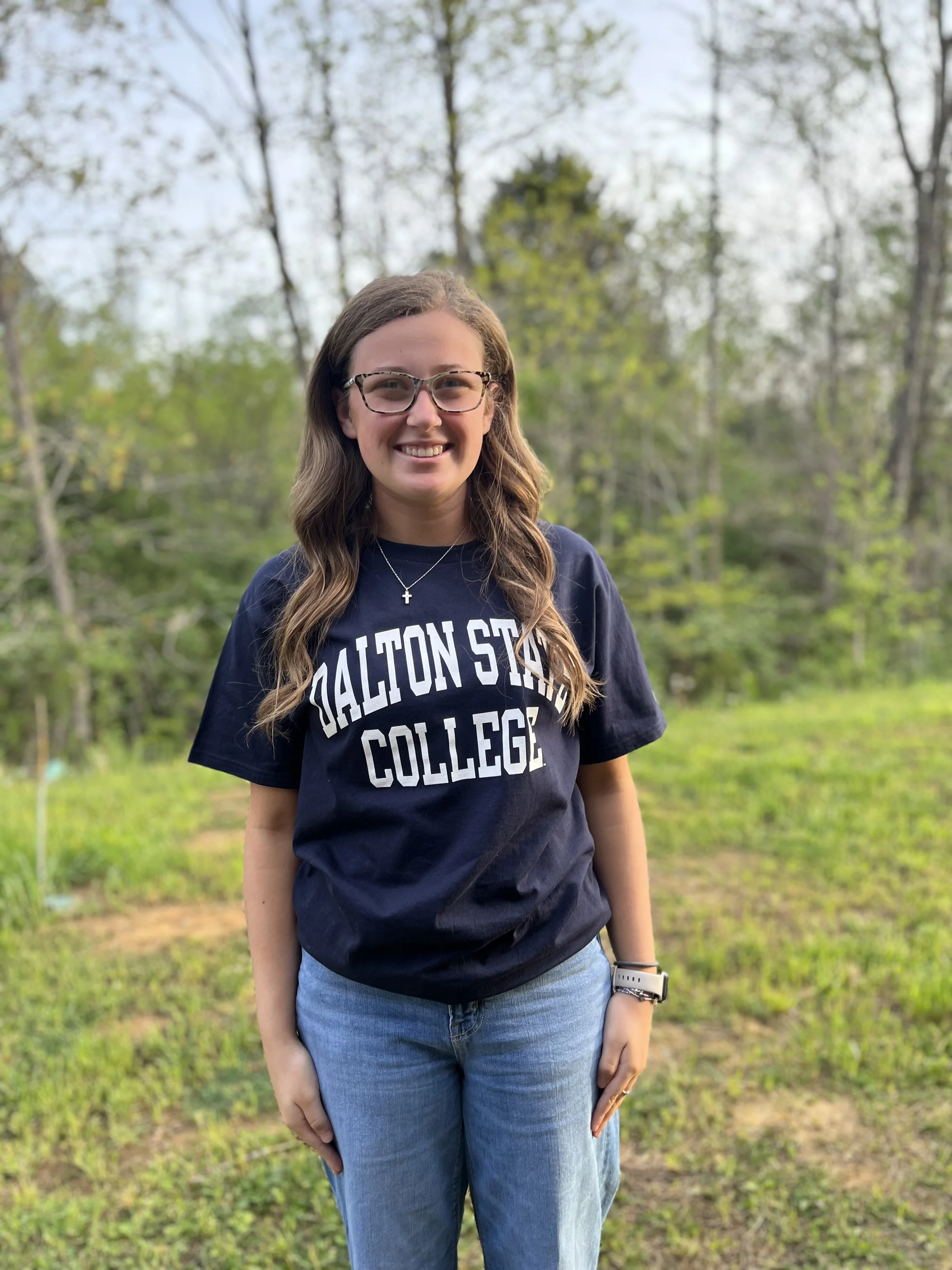 A young woman with glasses smiling at the camera, sitting at a table in a classroom or office setting.