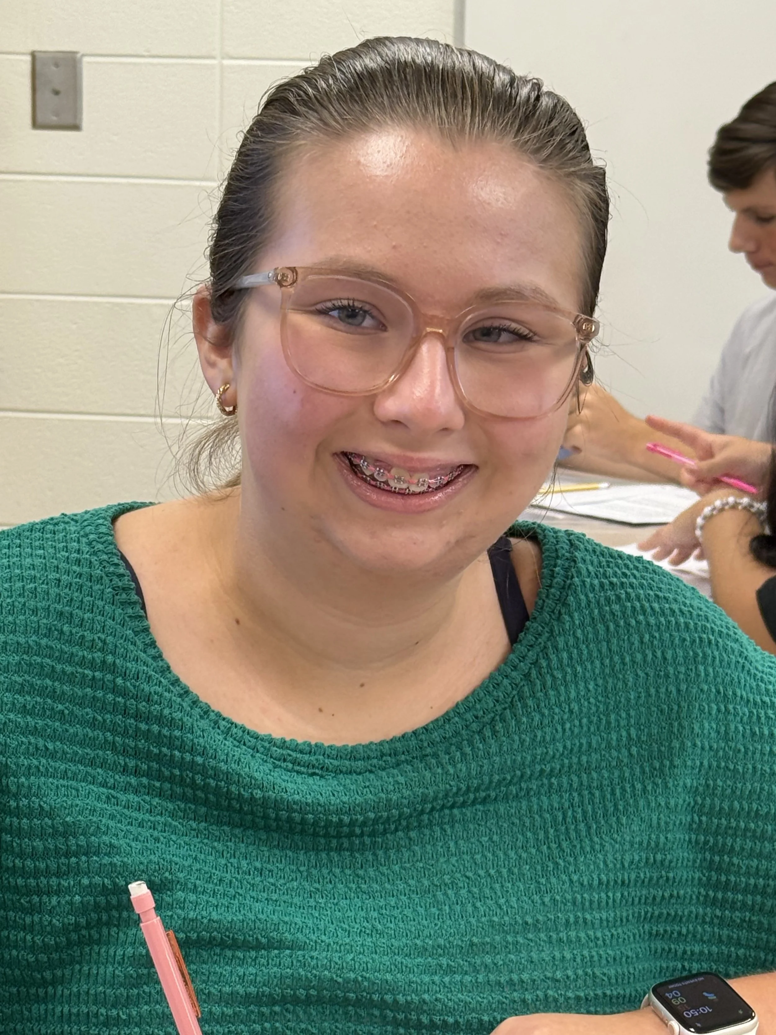 A young girl wearing glasses and a red shirt sitting at a table, resting her chin on her hand, looking directly at the camera. She has a smartwatch on her wrist and is in a room with other people in the background.