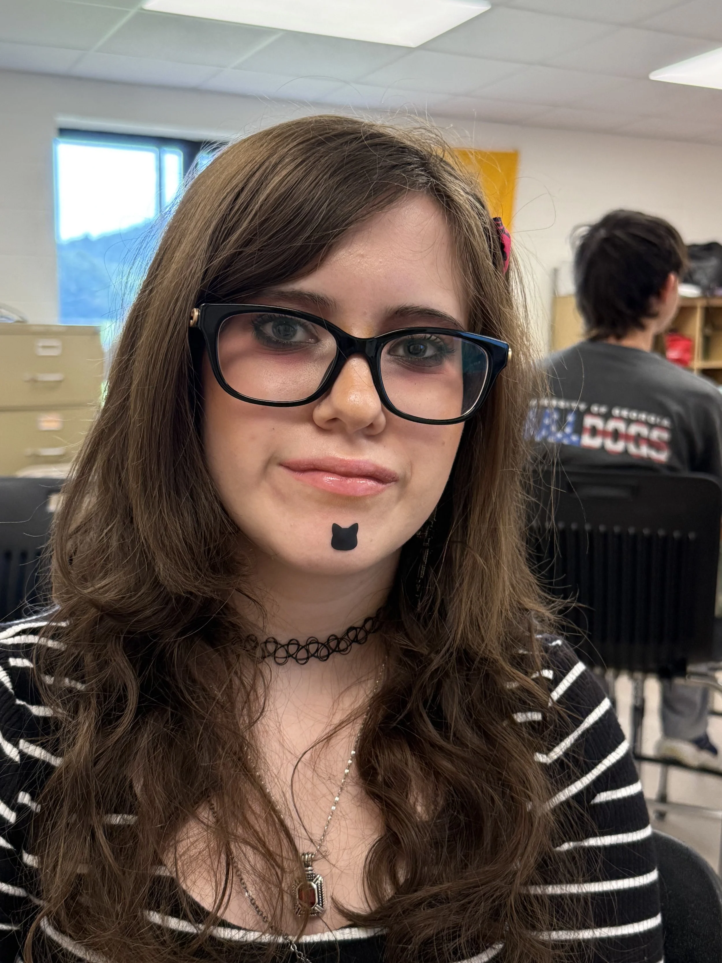 A young woman with brown, curly hair and glasses, wearing a black shirt with white text and a black hoodie, and jewelry including a choker and beaded necklace with a cross pendant, smiling slightly.