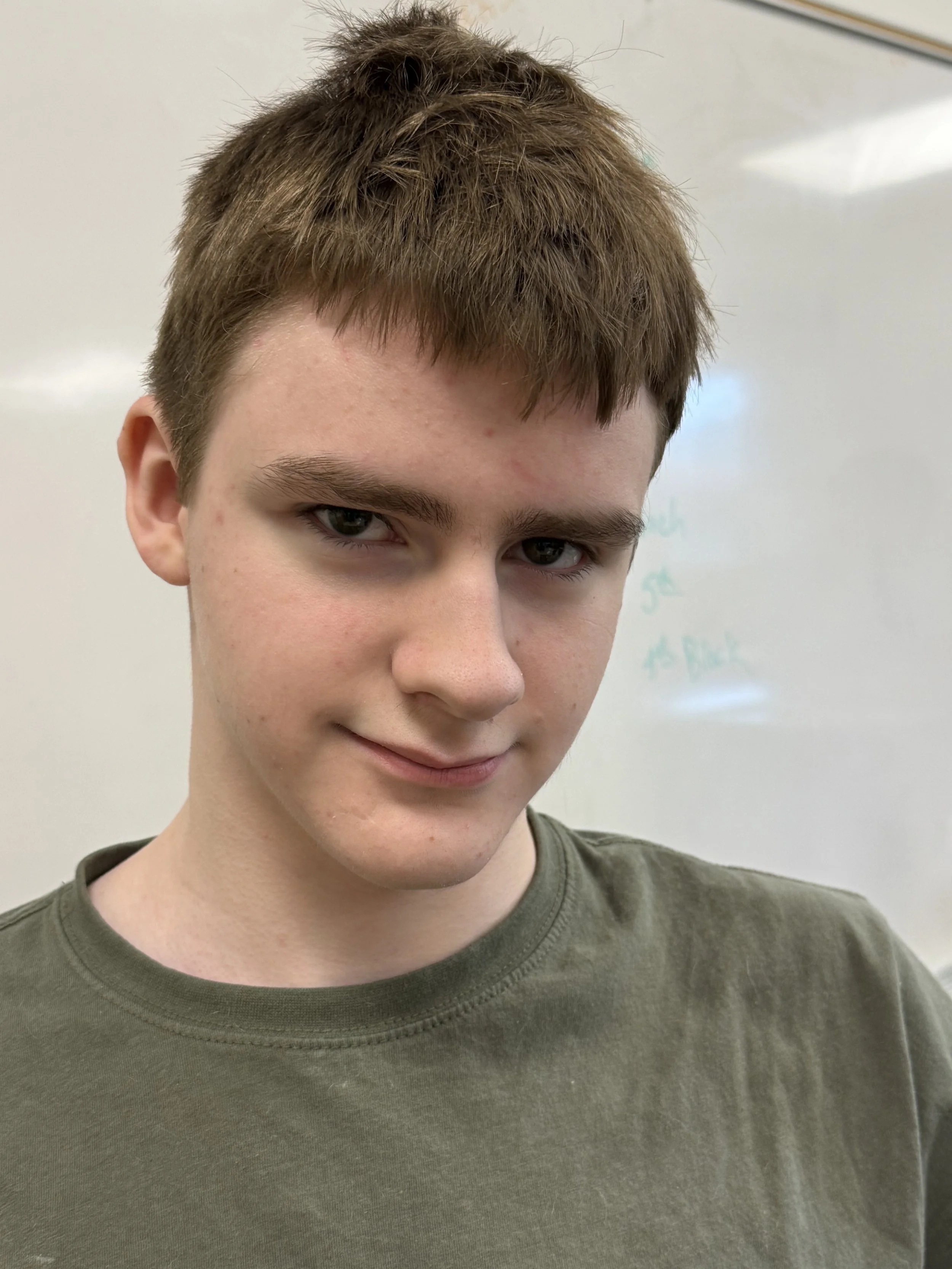 A smiling young boy with short brown hair and light skin standing in front of a whiteboard.