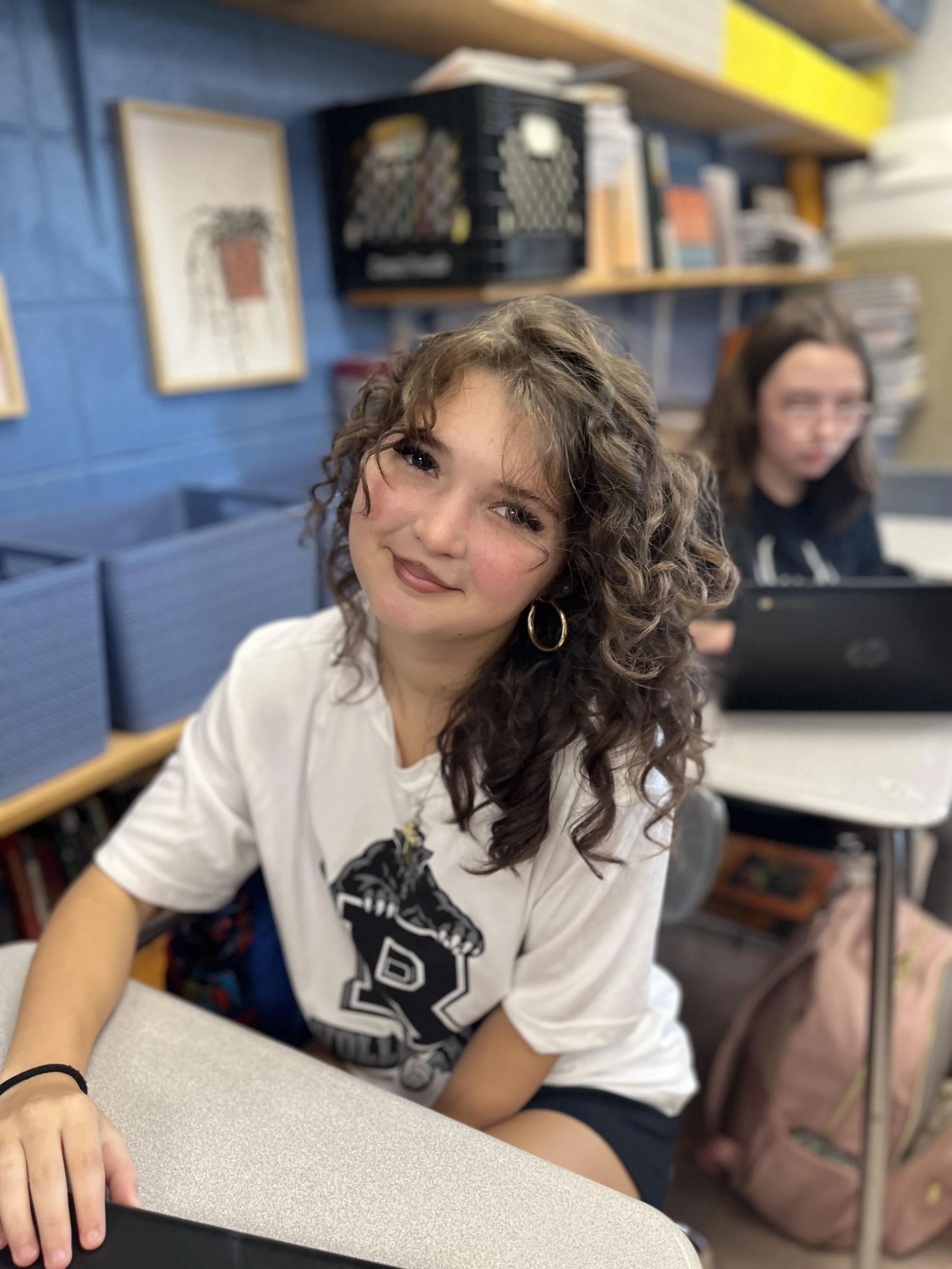 A young girl with a warm smile, sitting indoors, with her arms crossed on a table. She has dark braided hair and is wearing a plaid dress with ruffled sleeves, with a background that appears to be a classroom or similar setting.