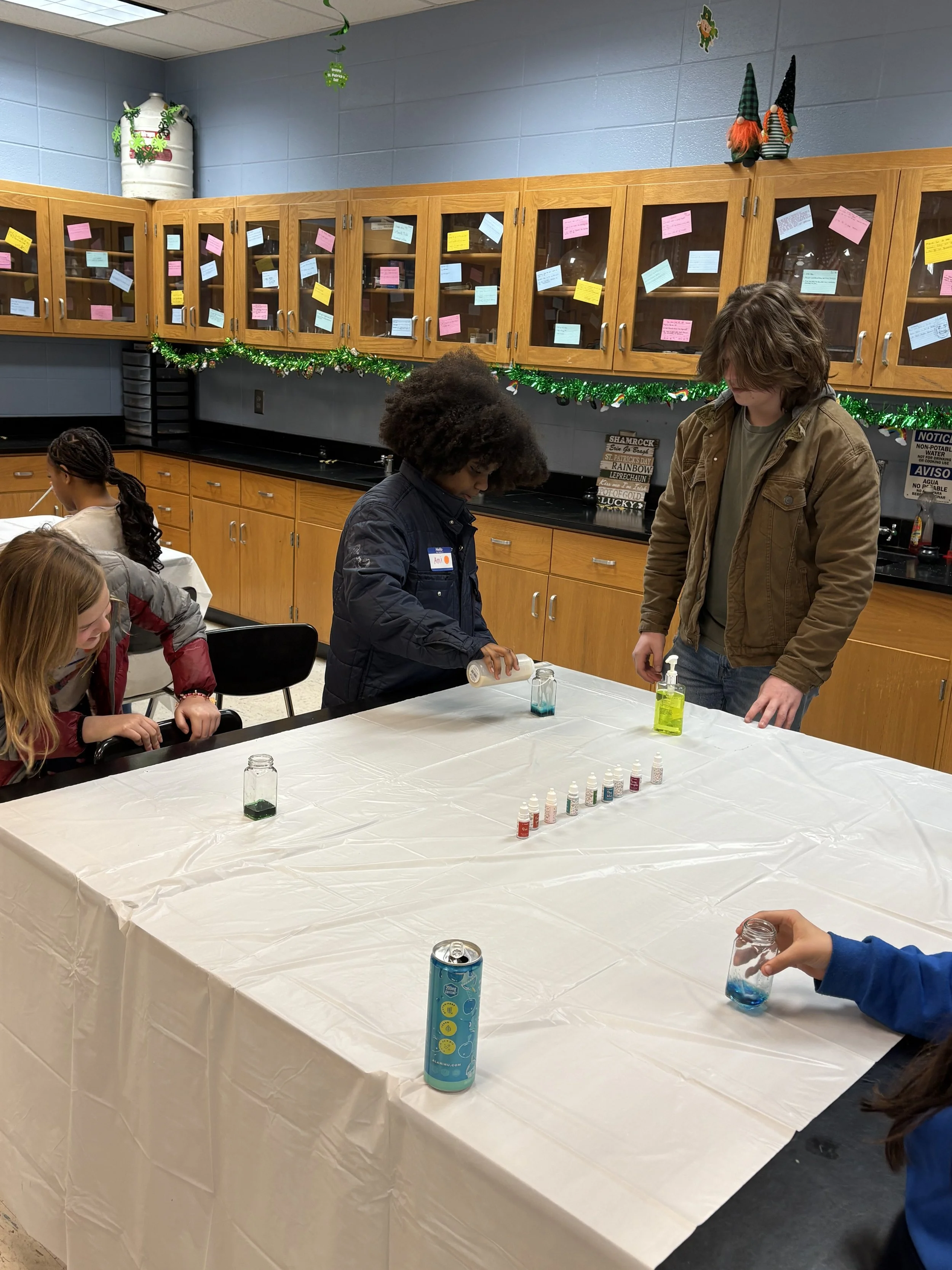 Children participating in a science activity at a school science lab. A girl is pouring a liquid into a jar, while other children observe. The table is covered with a white plastic tablecloth, and there are small bottles of colorful liquids arranged 