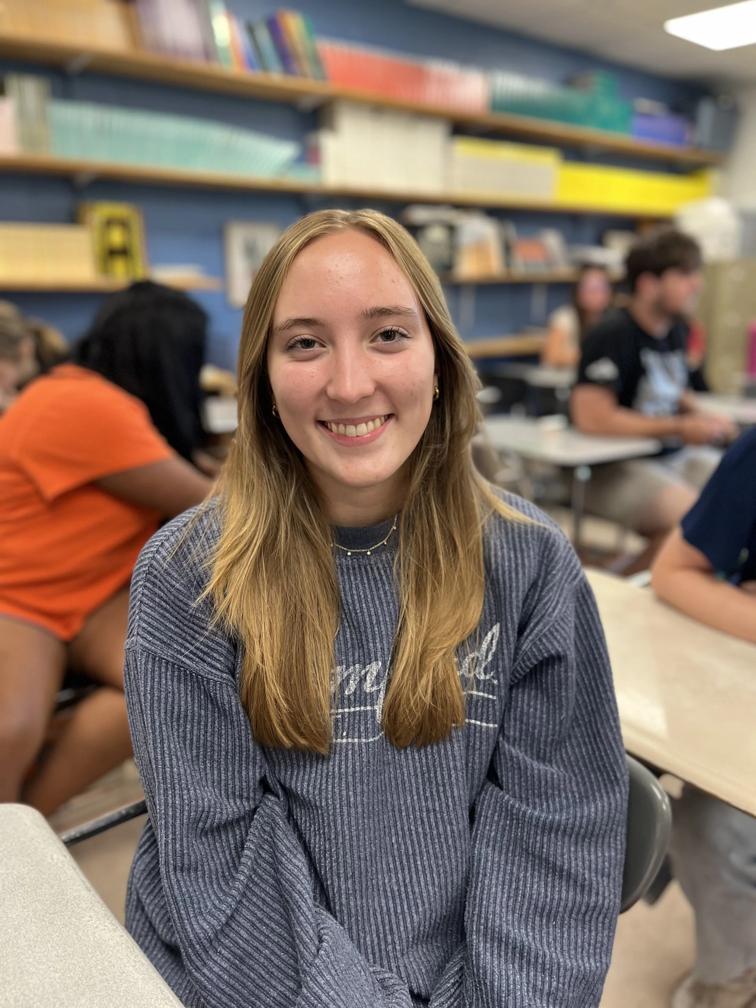Young girl with long red hair smiling at camera in classroom setting with students in background.