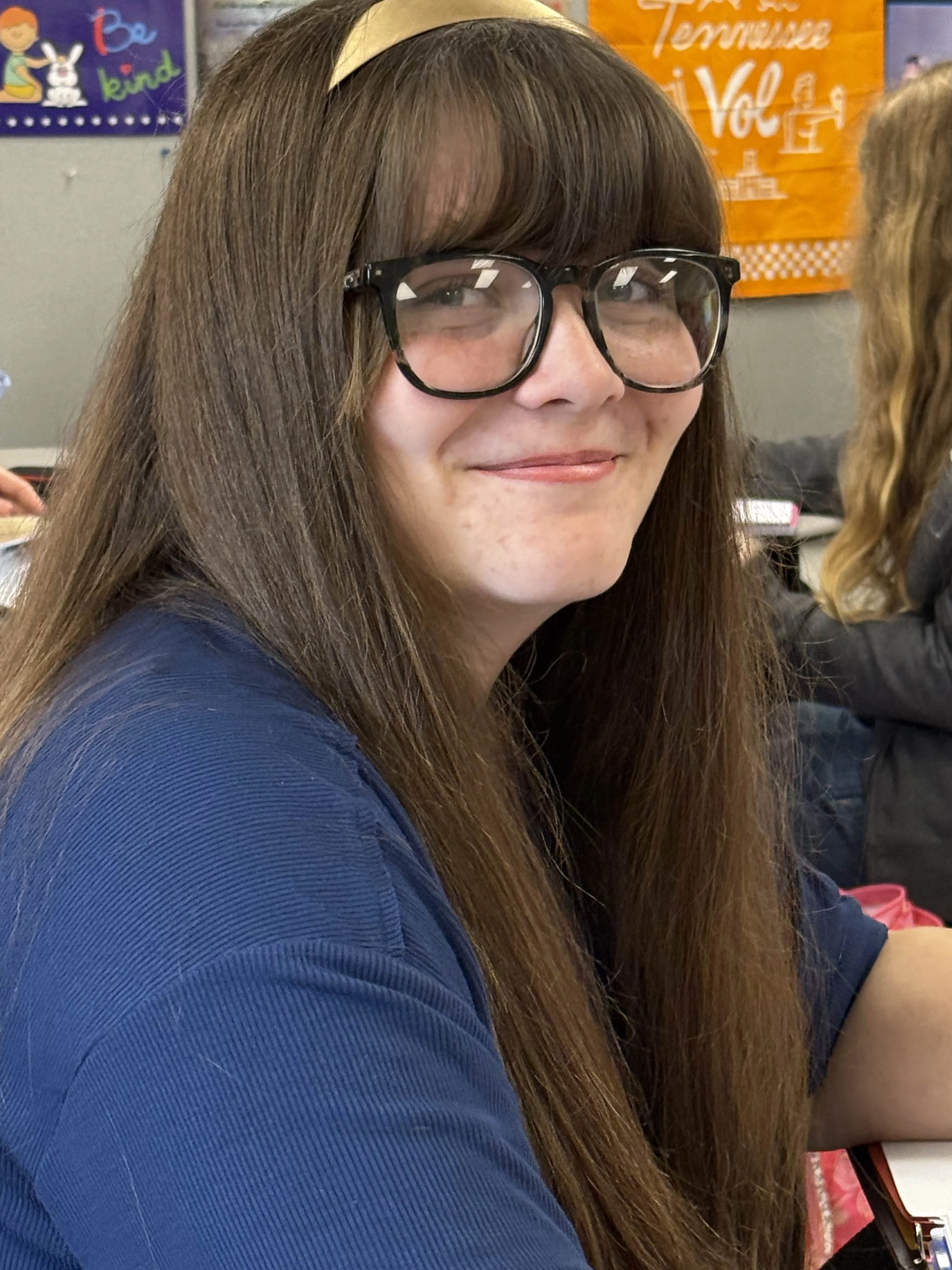 Smiling girl with long brown hair, black glasses, and a black shirt standing in front of a world map in a classroom.