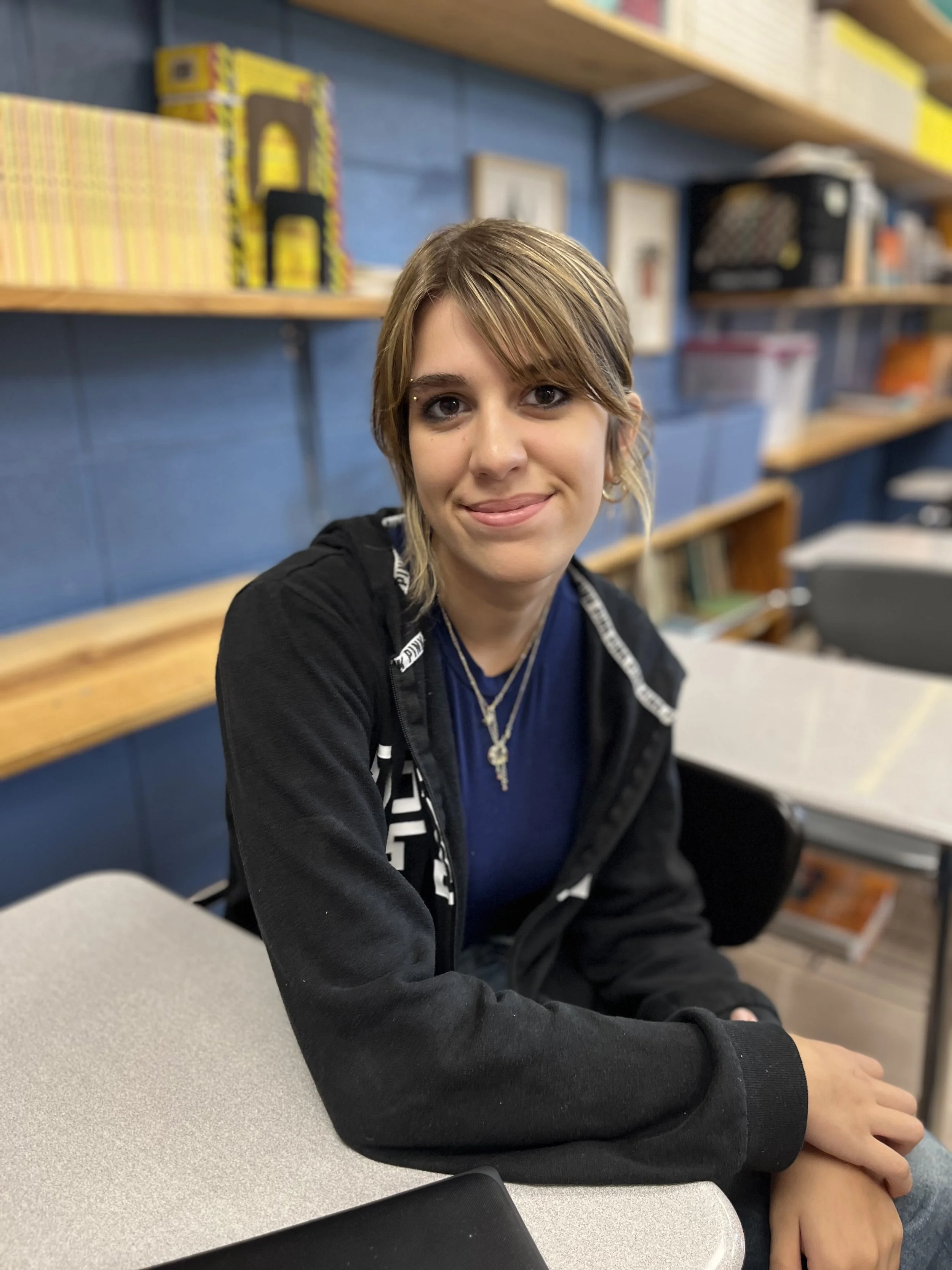 A young woman with blonde hair and blue eyes smiling at the camera, wearing a black t-shirt with shoulder cut-outs, sitting in a classroom with wooden shelves and books in the background.