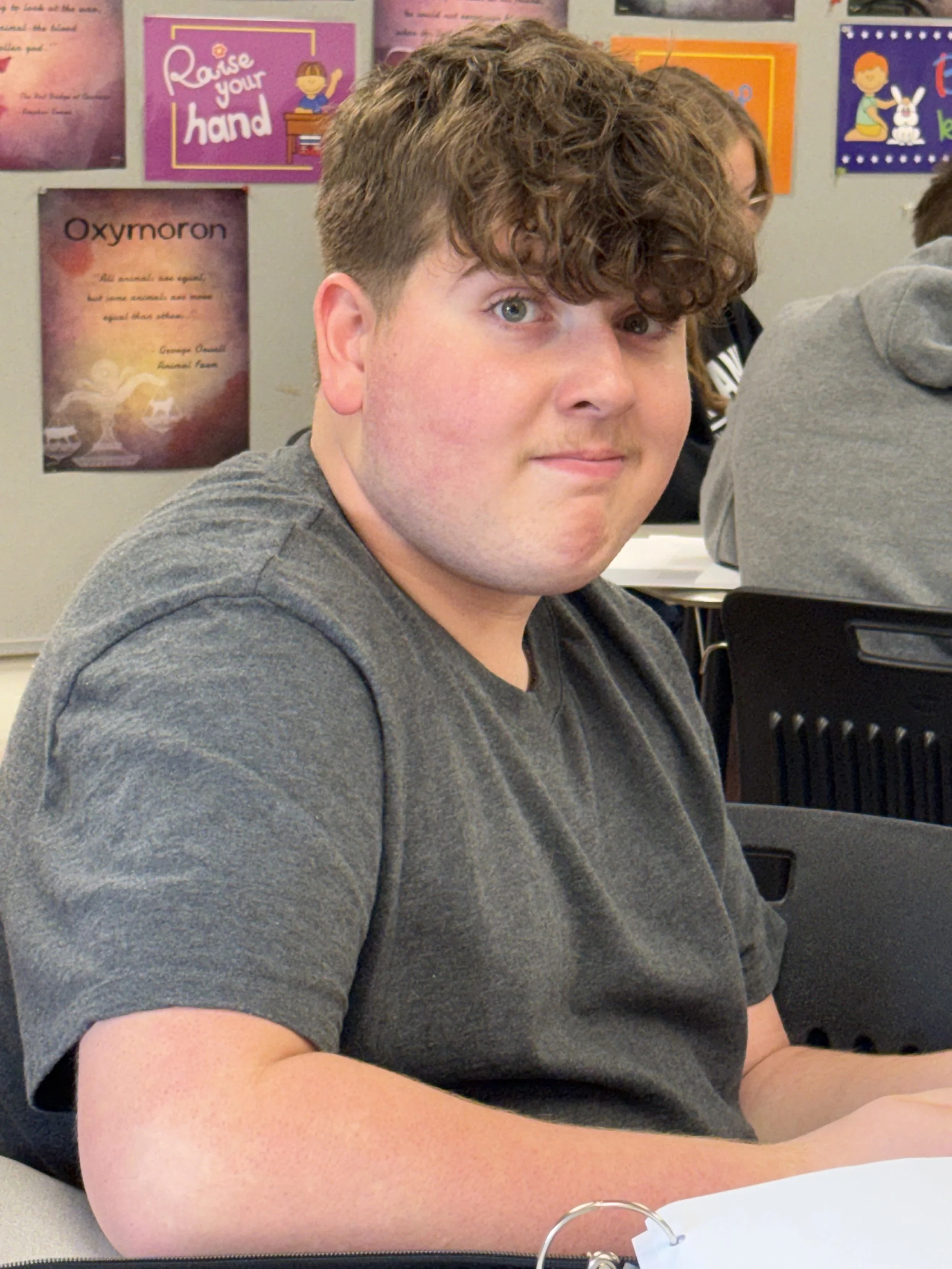 Young male student with curly hair wearing a green T-shirt, sitting in a classroom or library with shelves of books in the background.