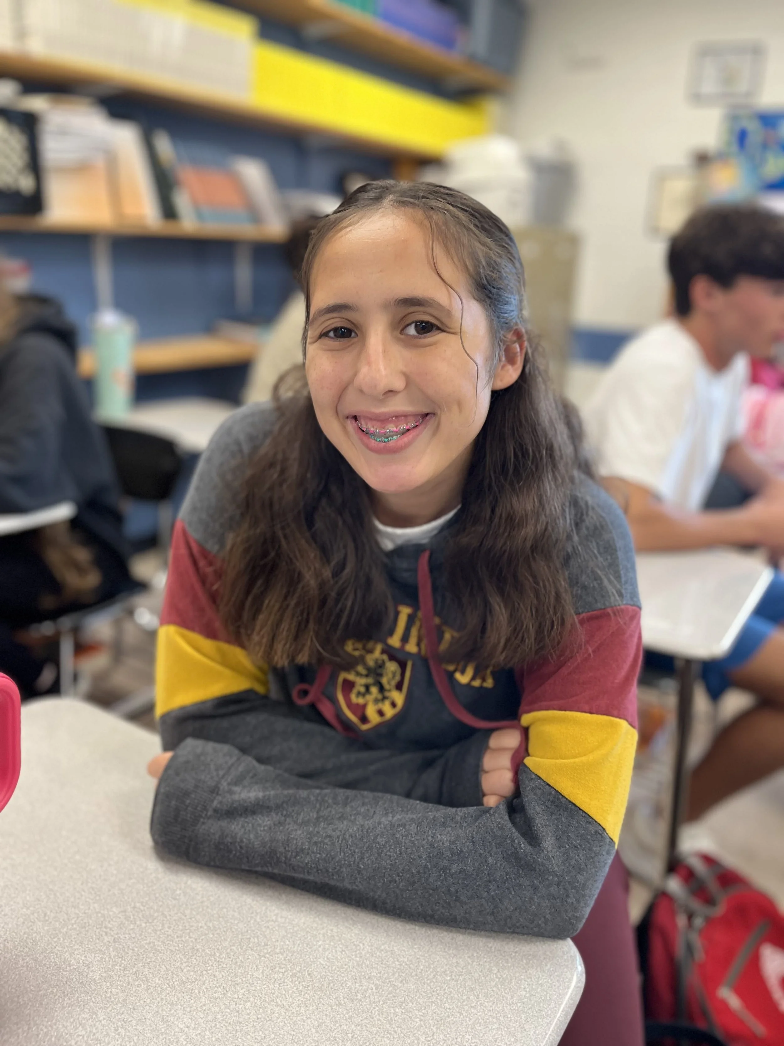 A young girl with long curly hair, braces, earrings, wearing a black hoodie with a tiger logo, smiling at the camera, sitting at a desk with a water bottle and laptop in a room with boxes and a filing cabinet in the background.
