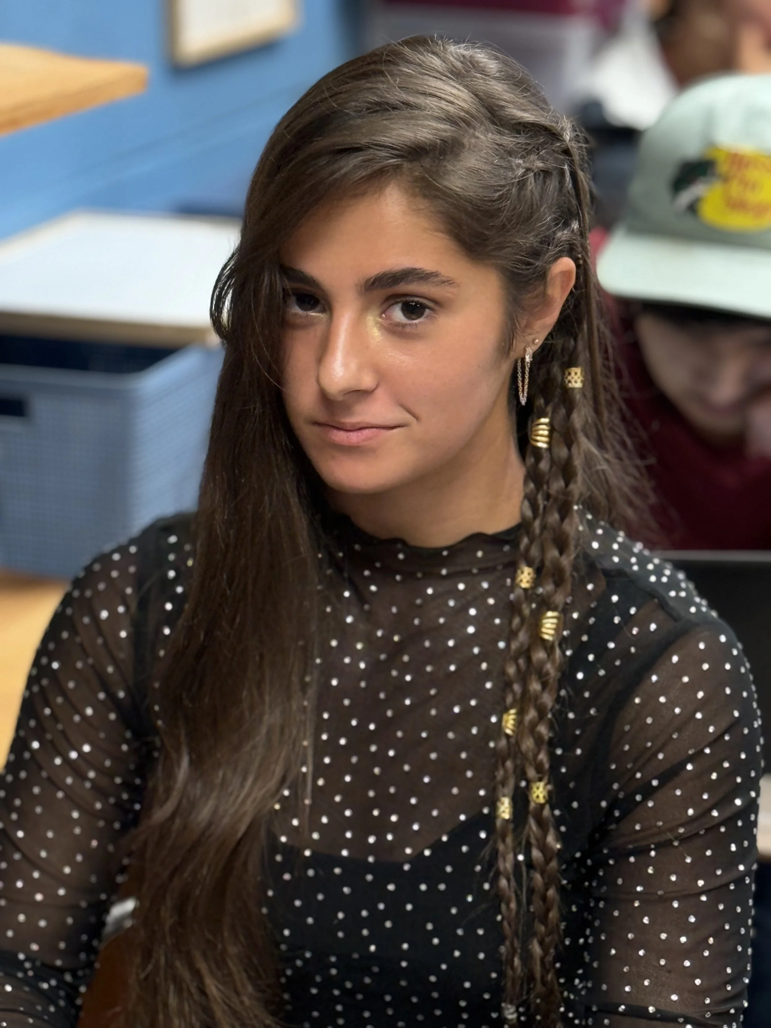 A young woman with long brown hair and a braid, wearing a black sheer top with white polka dots, sitting in a classroom setting.