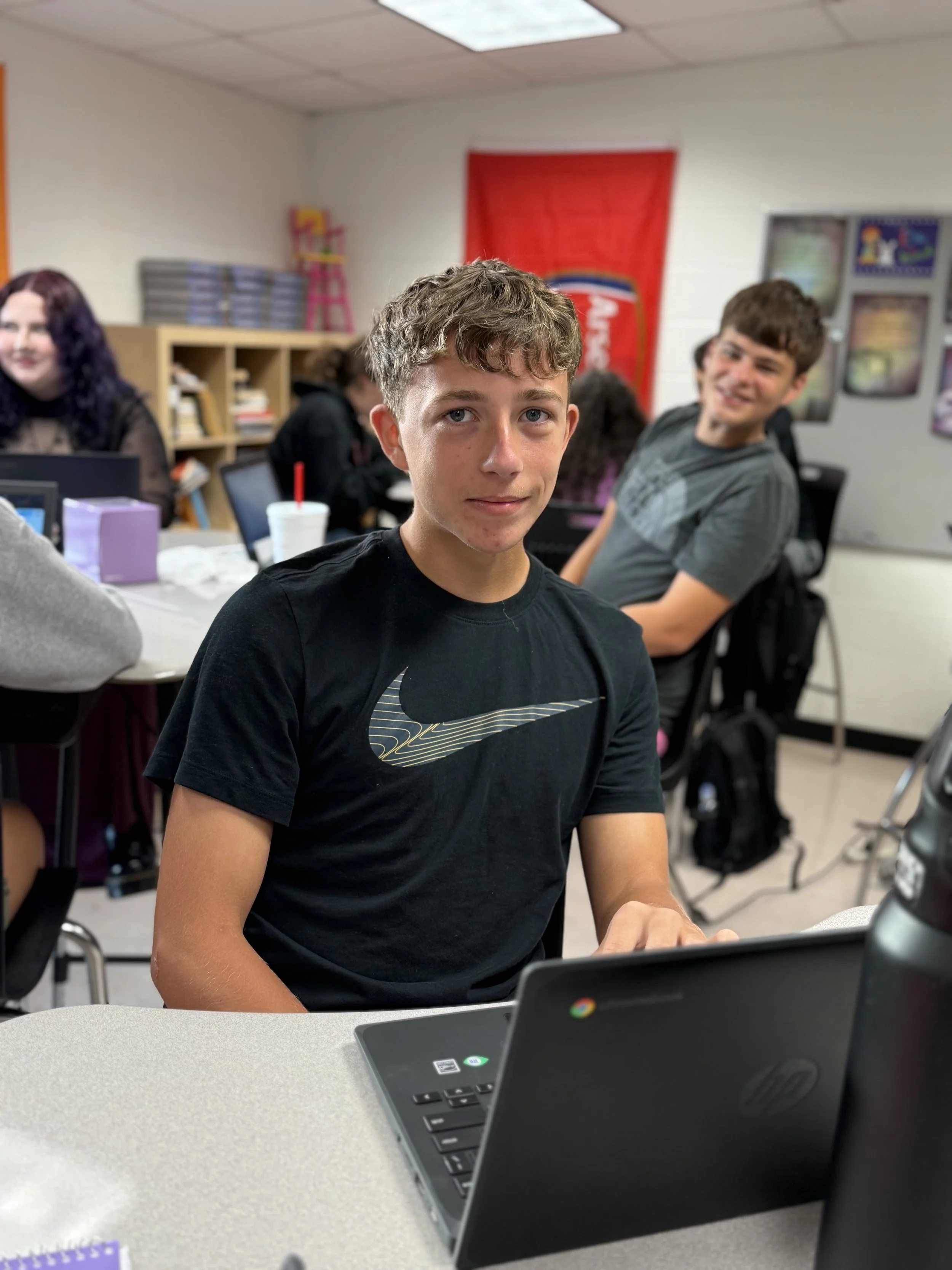 A boy sitting at a table with a laptop in a classroom, with other students and a teacher visible in the background.