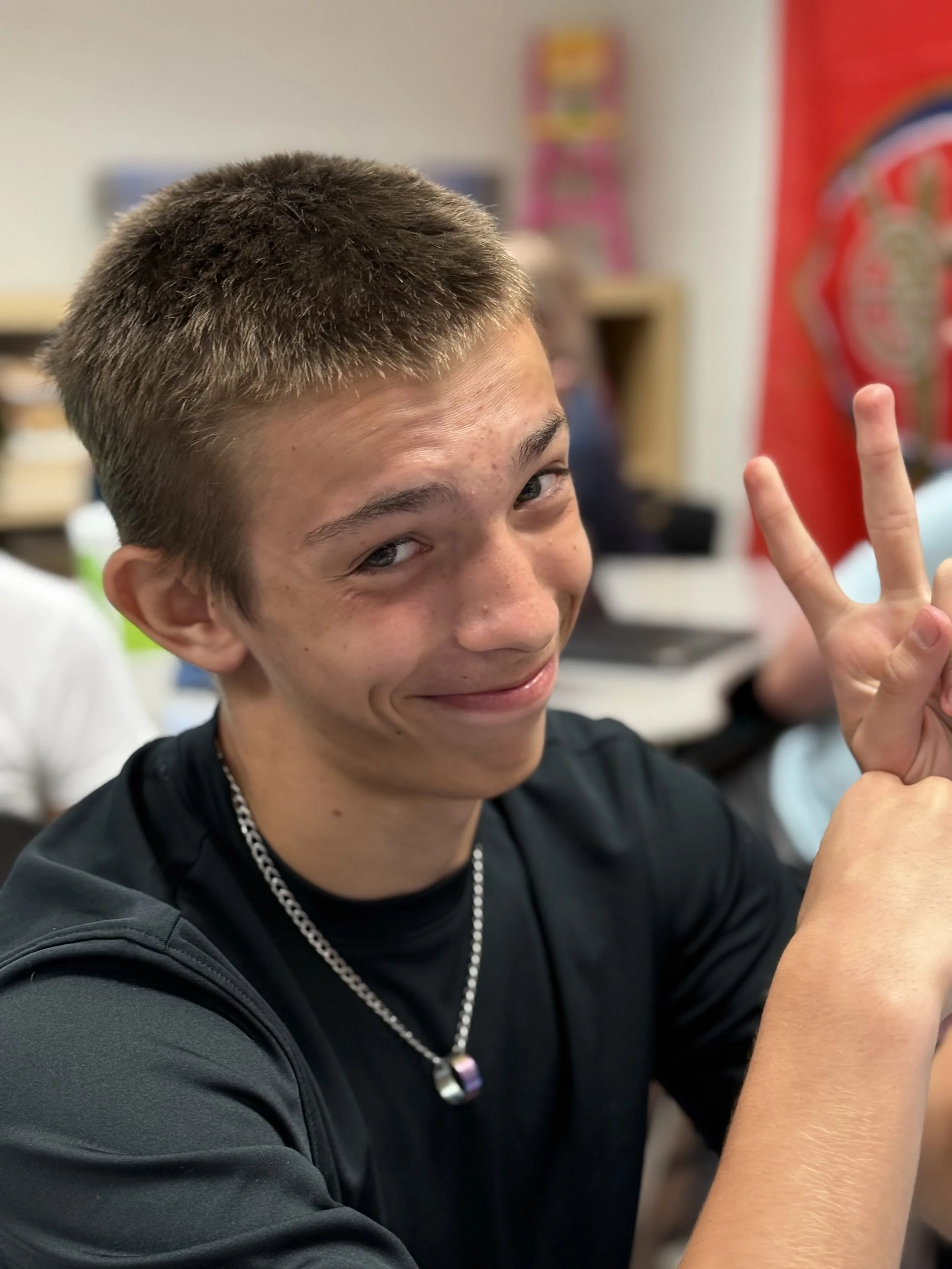 A teenage boy with short brown hair smiling and making a peace sign with his right hand in a classroom.