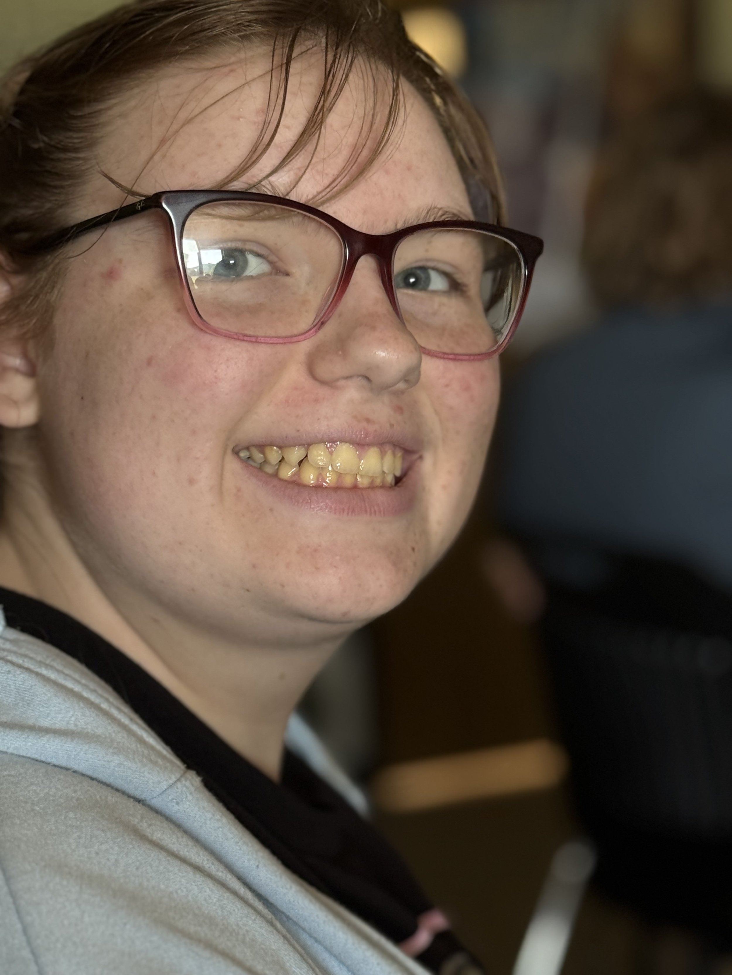 Close-up of a smiling woman with glasses and red hair, showing her teeth, with face freckles and short hair, indoors.
