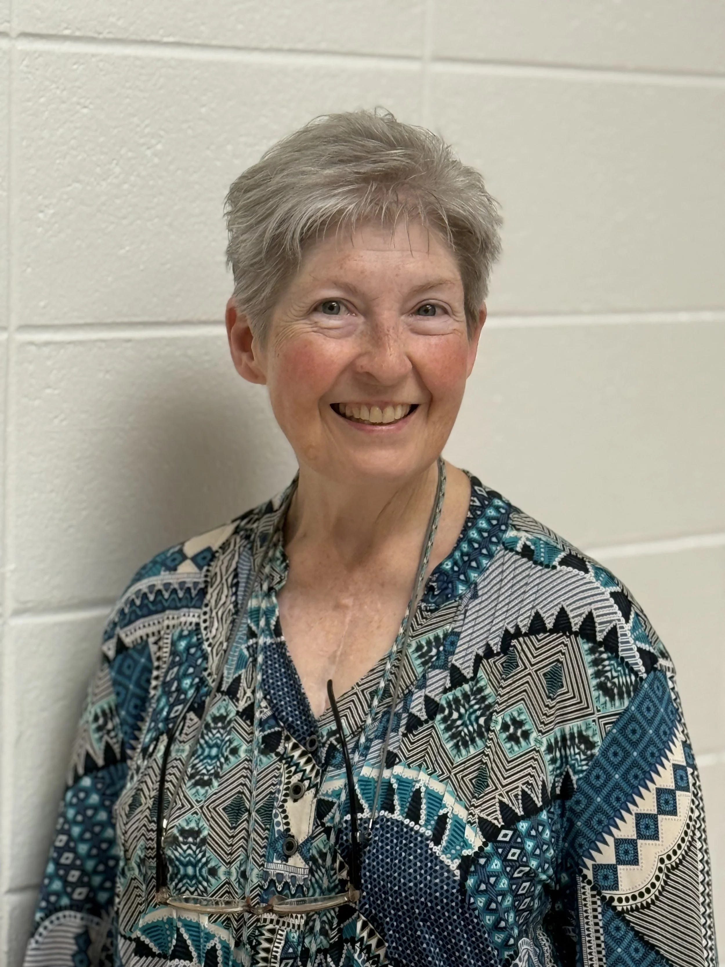 A smiling older woman with short gray hair, wearing a patterned blue, black, and white blouse, standing against a white cinder block wall.
