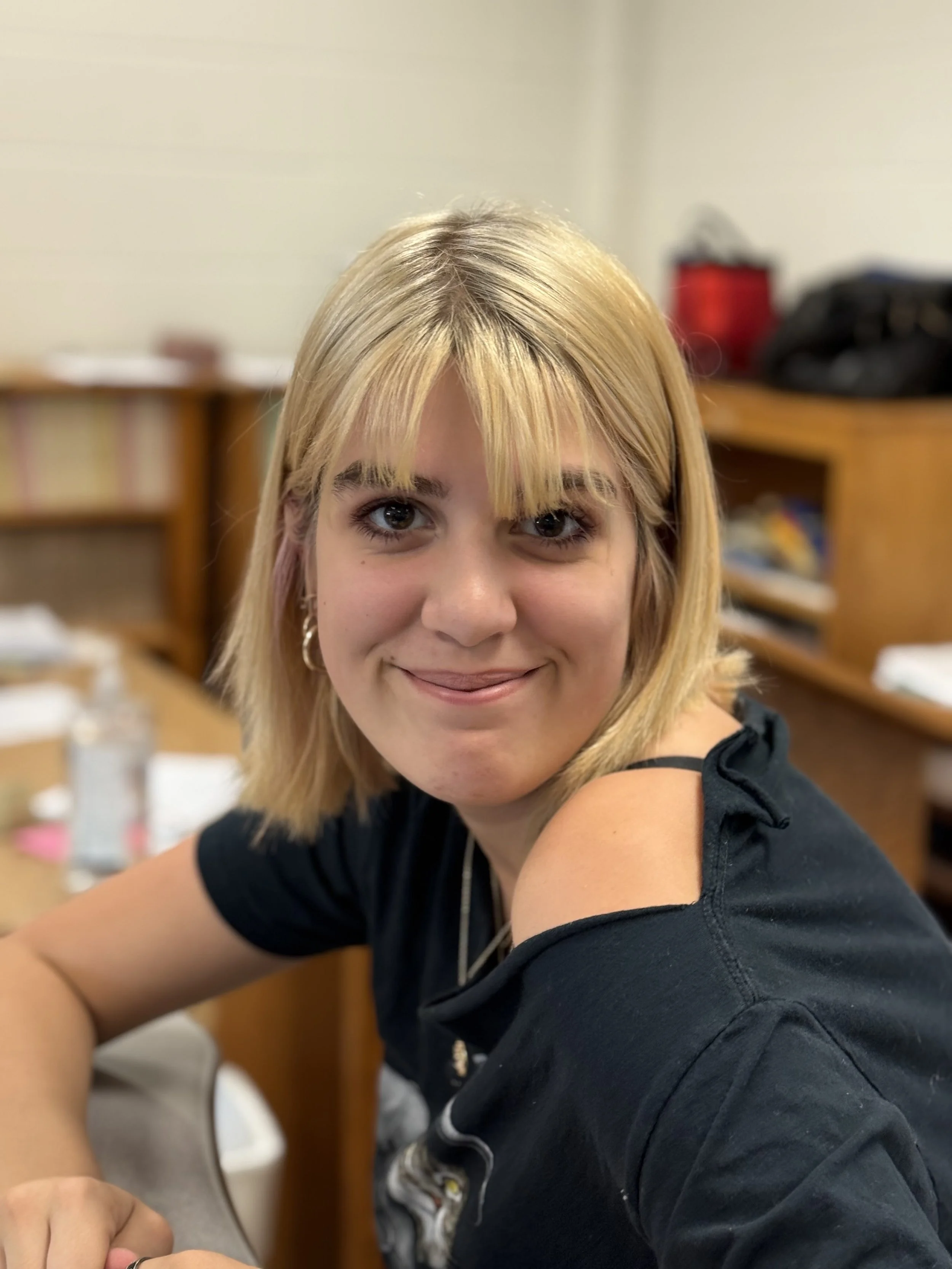 A young woman with blonde hair and blue eyes smiling at the camera, wearing a black t-shirt with shoulder cut-outs, sitting in a classroom with wooden shelves and books in the background.